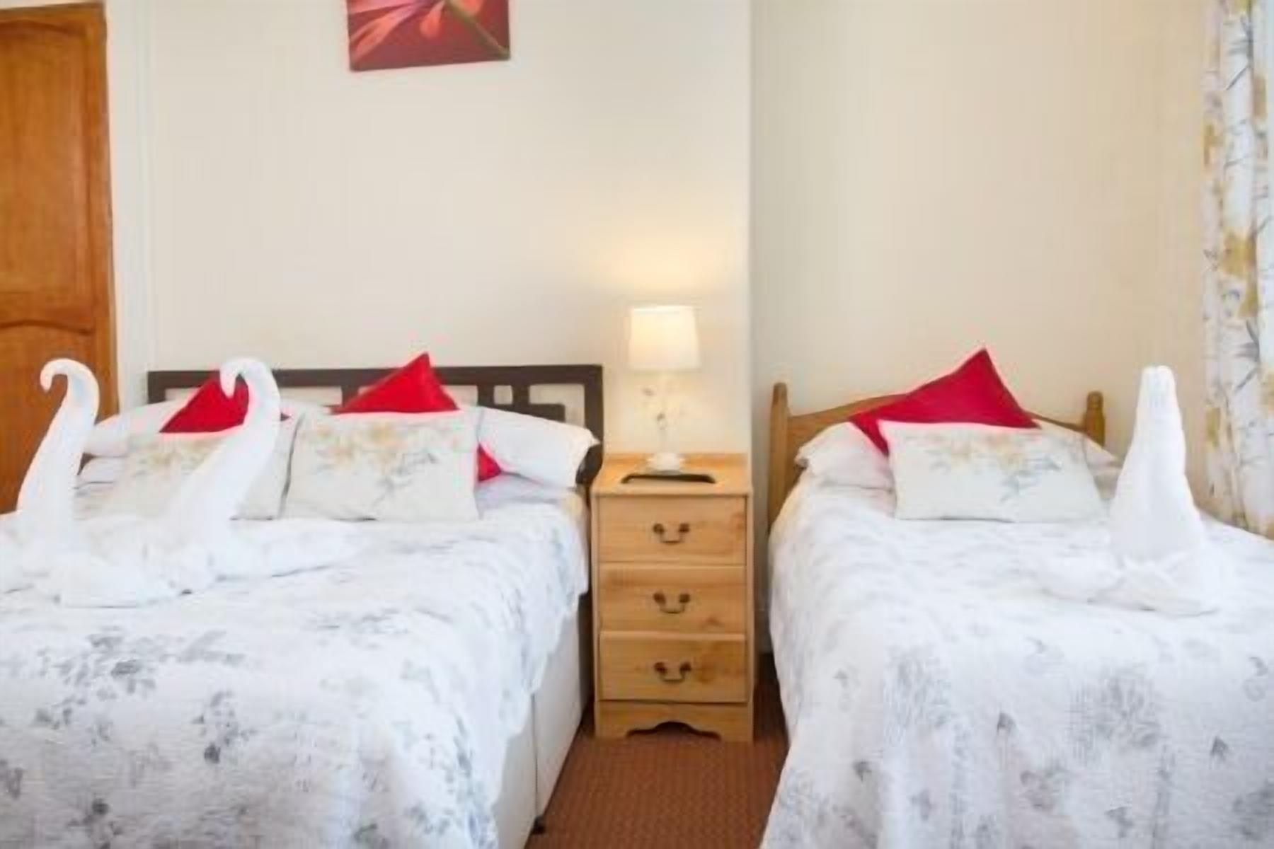 Een hotelkamer met twee bedden, elk met een wit bloemenbedekking en rode kussens, een houten nachtkastje met een lamp, en een houten deur.