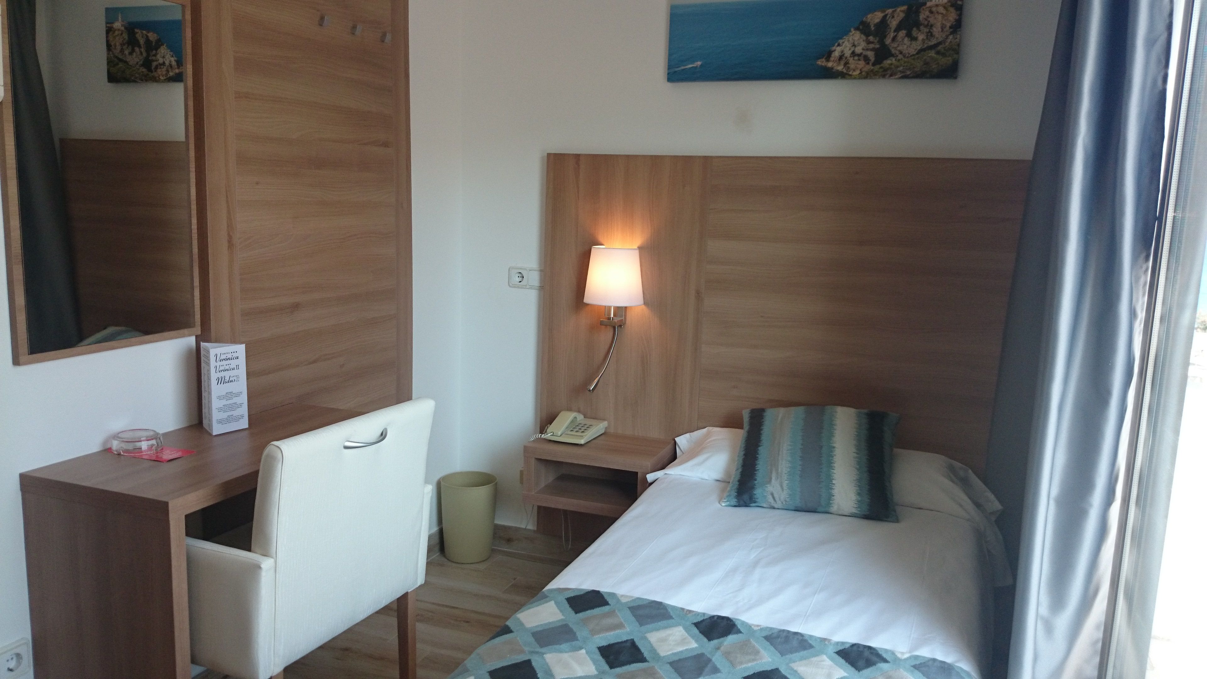 Habitación de hotel con cama, escritorio, silla, mesita de noche con lámpara, cesto de basura y espejo.