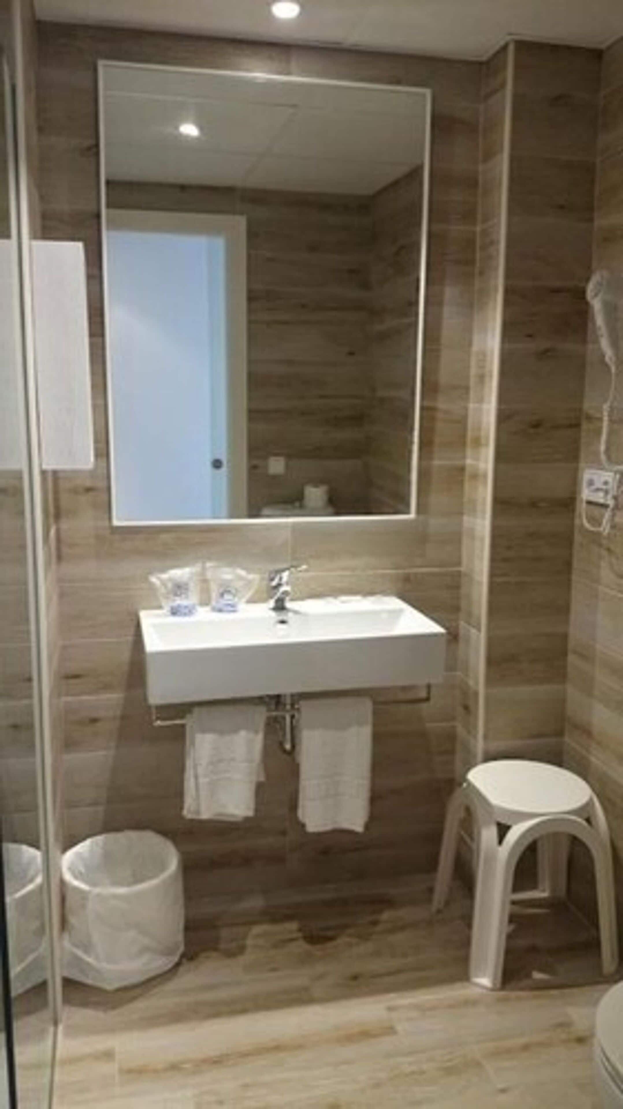 A bathroom with a white sink, mirror, and white stool. There is a trash can and a towel rack.