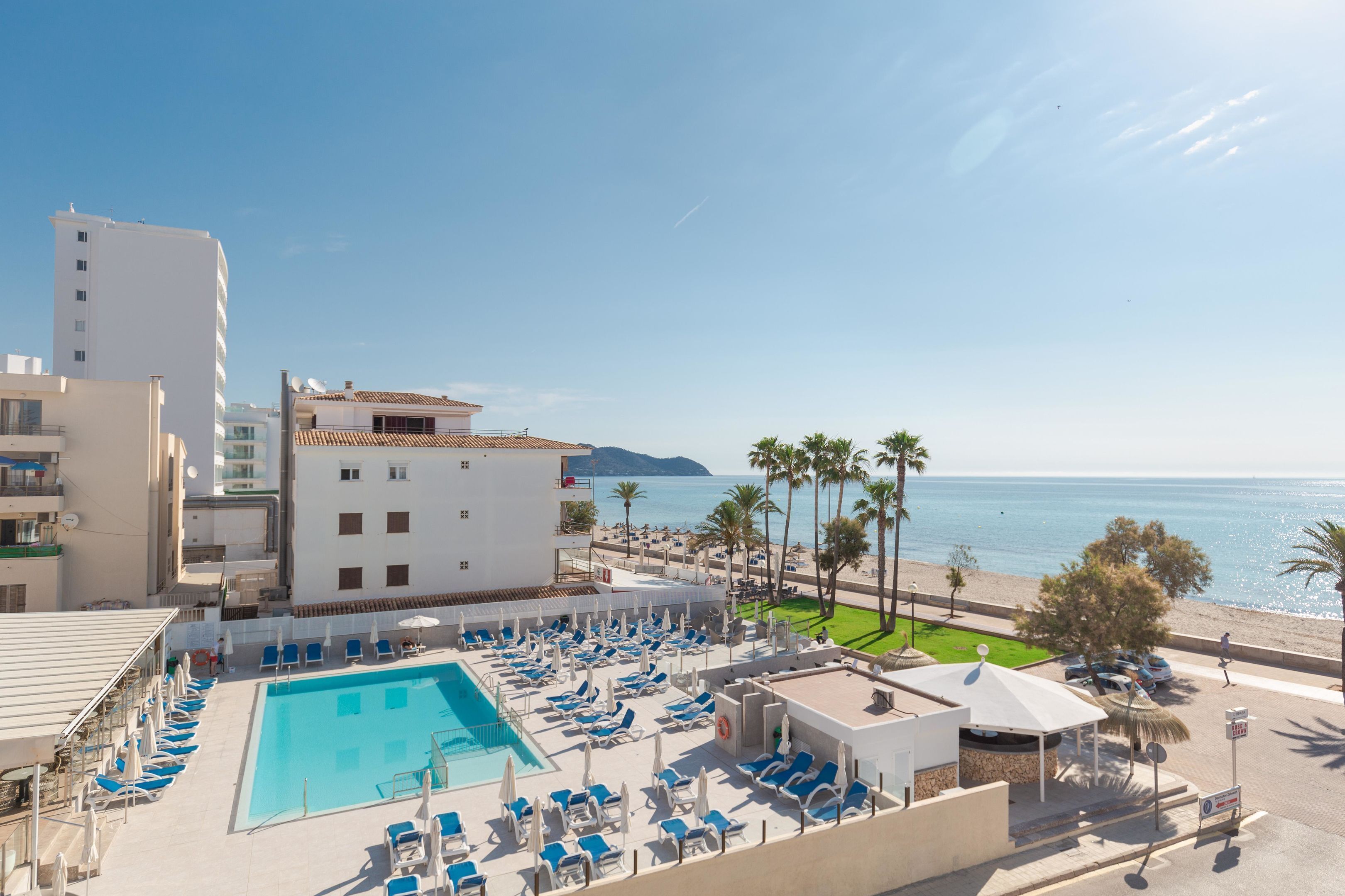 Un área de piscina en la azotea con sillas reclinables azules y sombrillas, una vista despejada del mar y un edificio con tejado de baldosas.