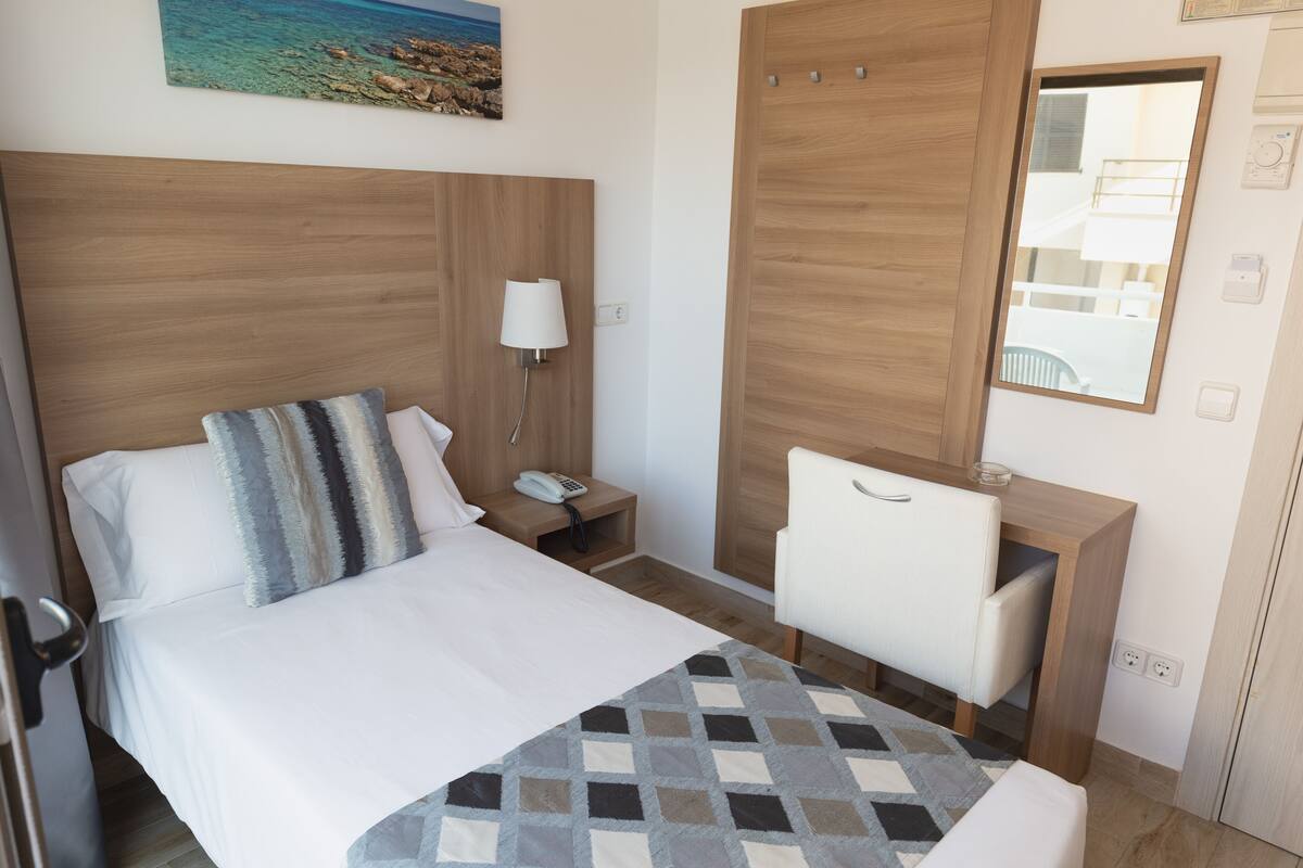 A hotel room with a bed, bedside table with a lamp, a desk with a chair, a mirror, and a painting on the wall.