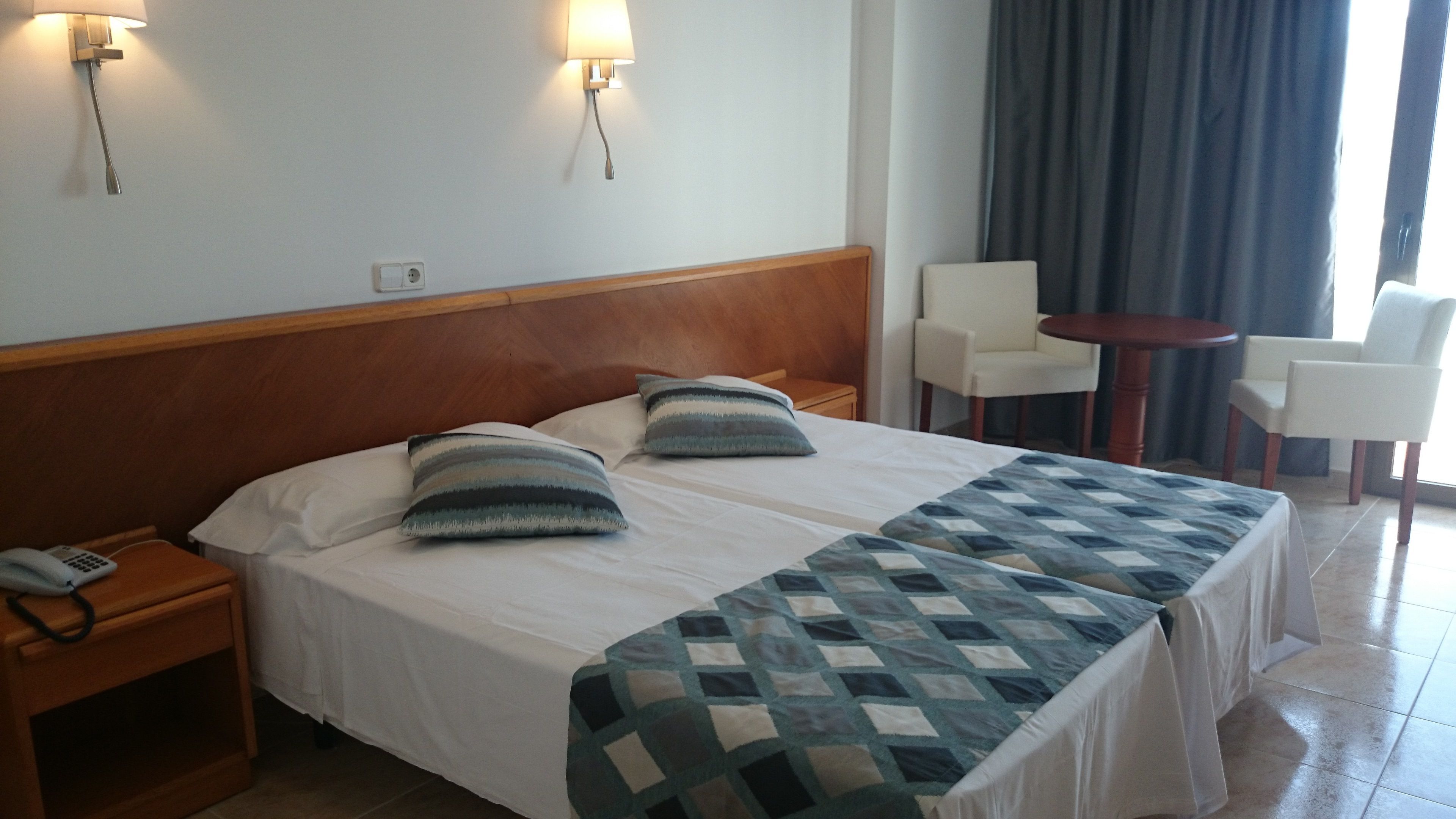 A hotel room with a large bed, two bedside tables, a chair, a small table, and a window with curtains.