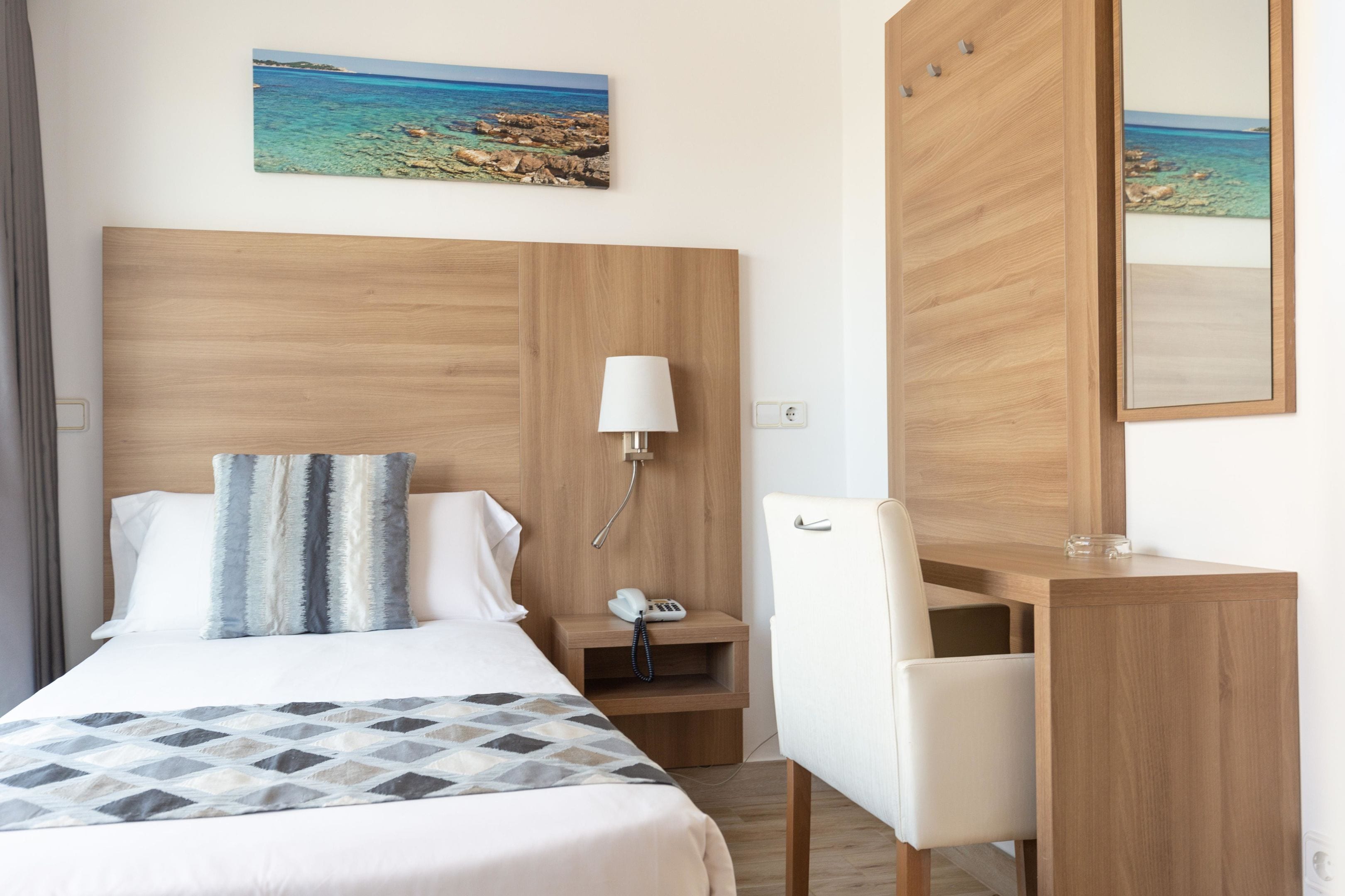 Une chambre d’hôtel avec une tête de lit en bois, un lit recouvert d’une couverture à motifs, une table de chevet avec une lampe, un bureau avec une chaise et un grand miroir.