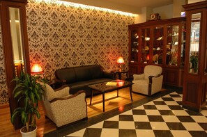 Una sala de estar con piso a cuadros, papel tapiz estampado, un sofá, dos sillones, una mesa de centro y un aparador con puertas de vidrio.