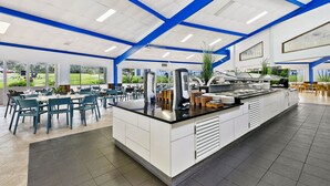 A spacious cafeteria with a central island, blue ceiling beams, and large windows offering views of the outdoors. - K'gari Beach Resort (Eurong)
