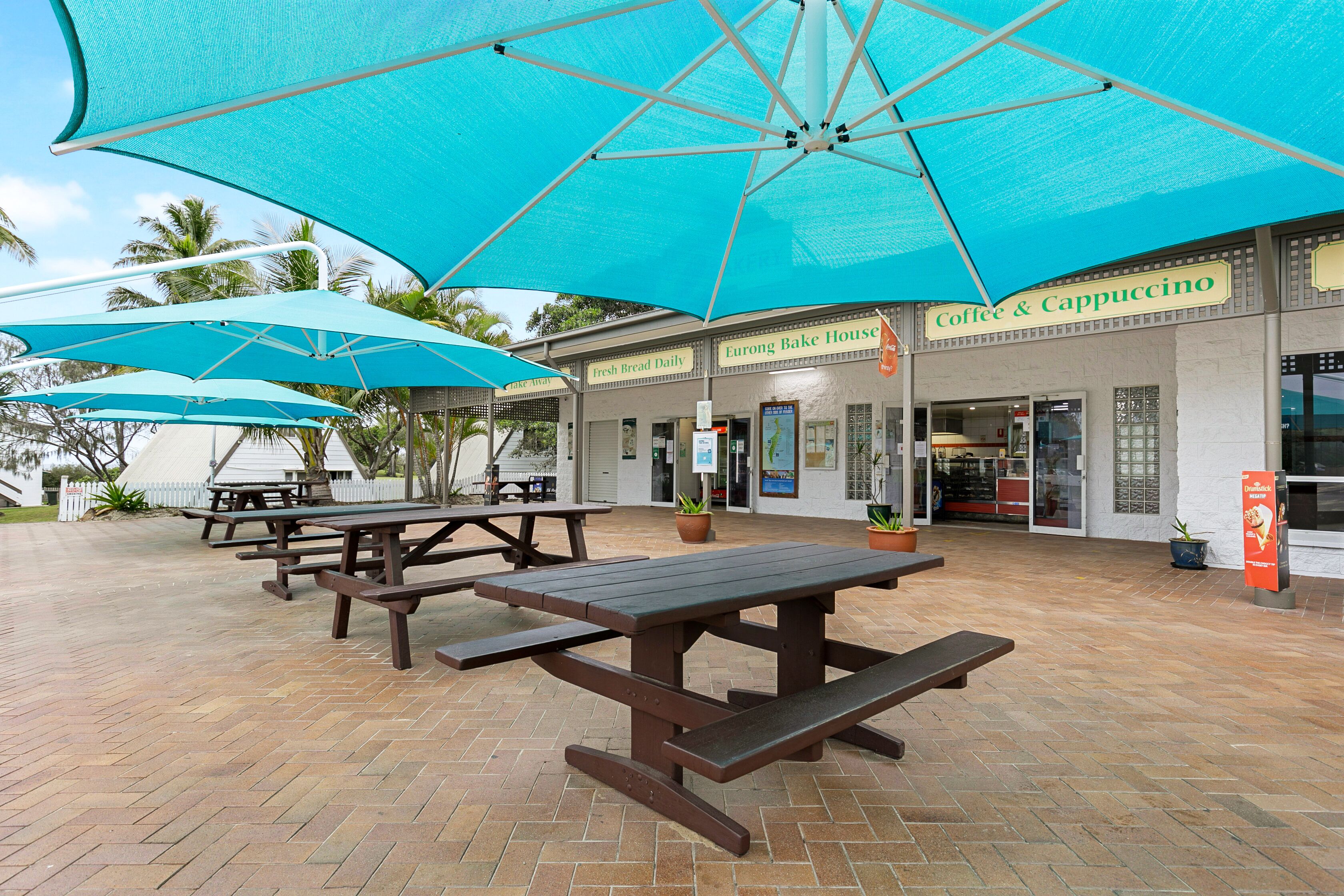Un espace extérieur aménagé avec des tables de pique-nique et des parasols, une boulangerie et un café.