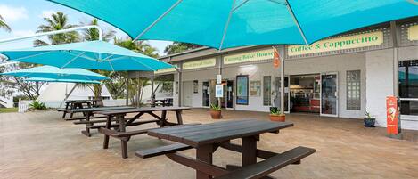 Un espace extérieur aménagé avec des tables de pique-nique et des parasols, une boulangerie et un café.
