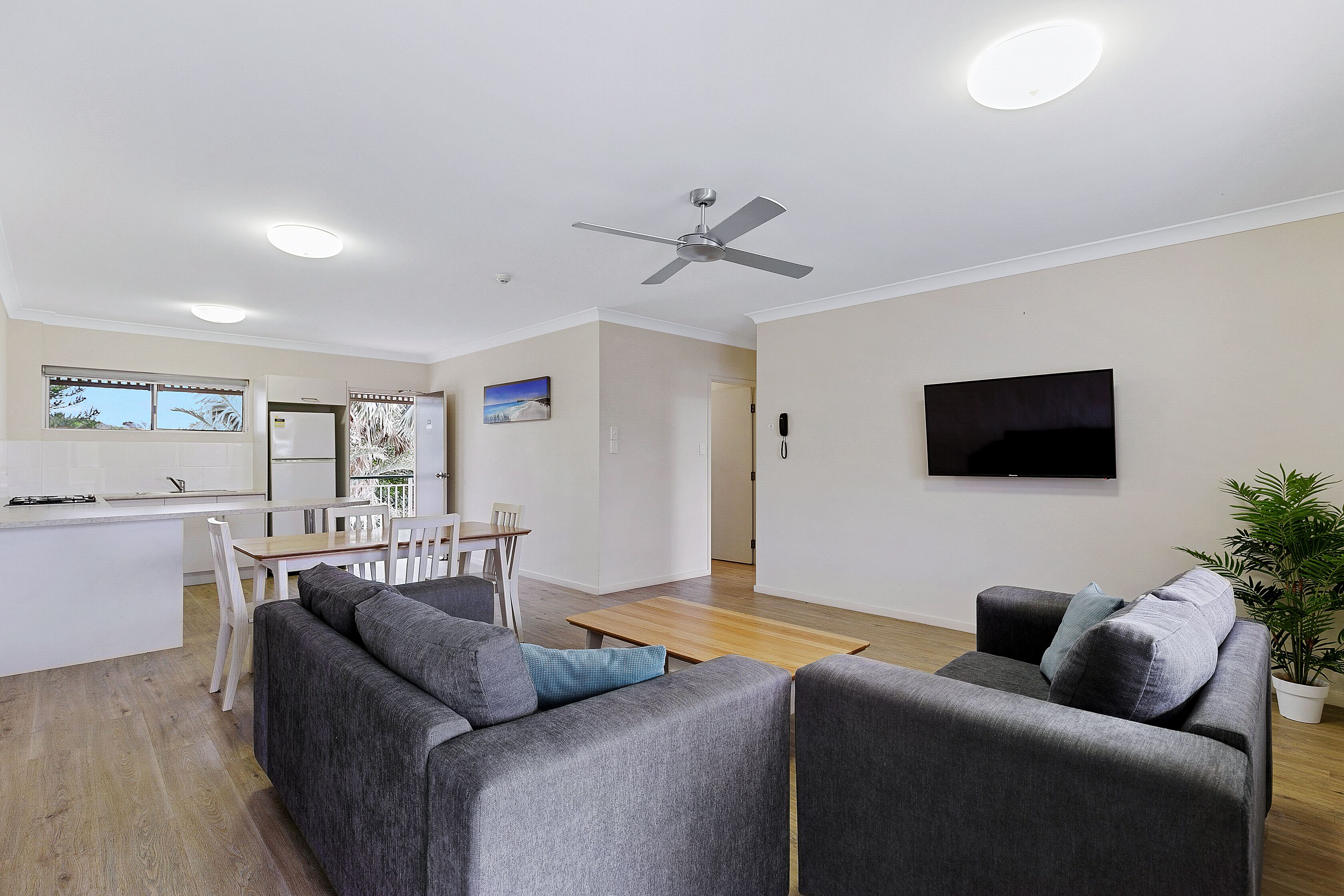 A modern living room with a grey sofa, a wooden dining table, and a ceiling fan.