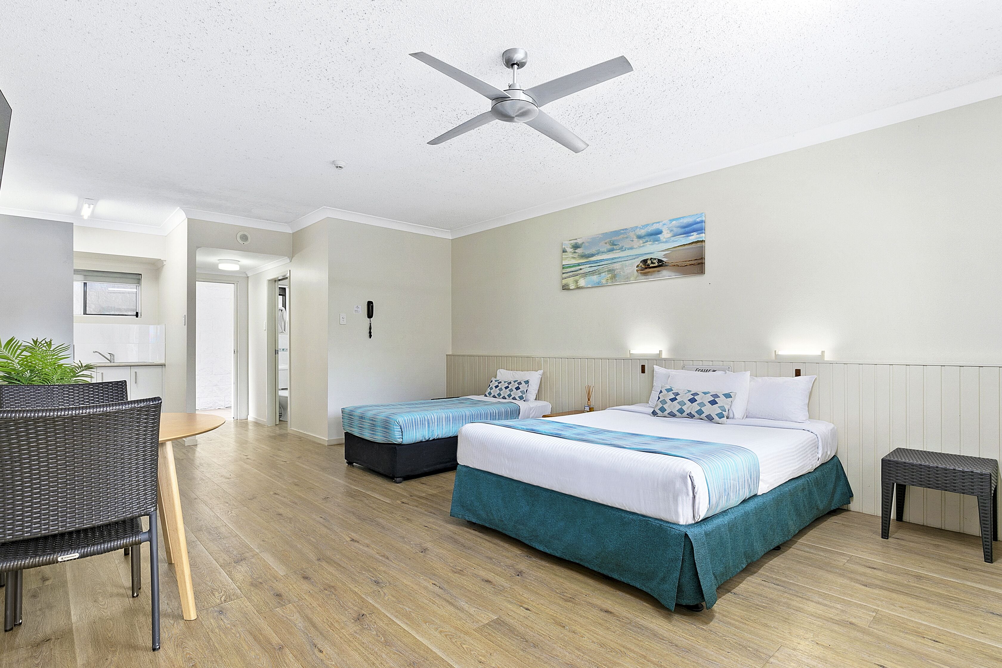 A hotel room with two beds, a dining area with a table and chairs, a ceiling fan, and a painting on the wall.