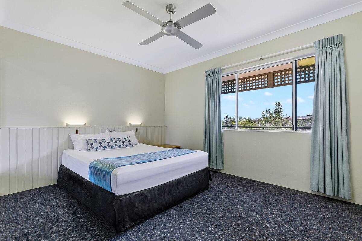 A hotel room with a bed, a ceiling fan, a window with curtains, and a view of the outside.
