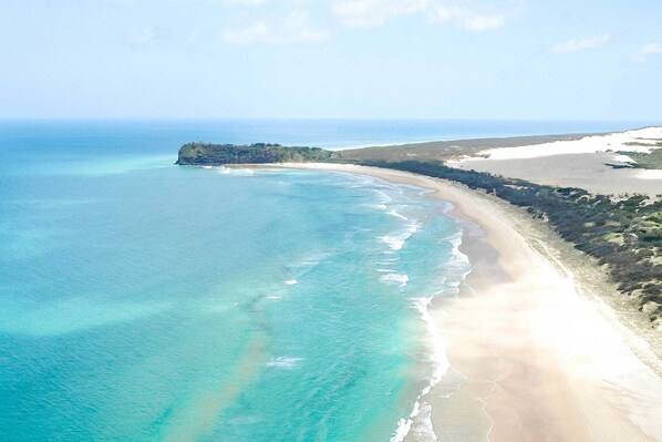 Vista aérea de una playa con aguas turquesas cristalinas, una orilla arenosa y una pequeña isla con vegetación.