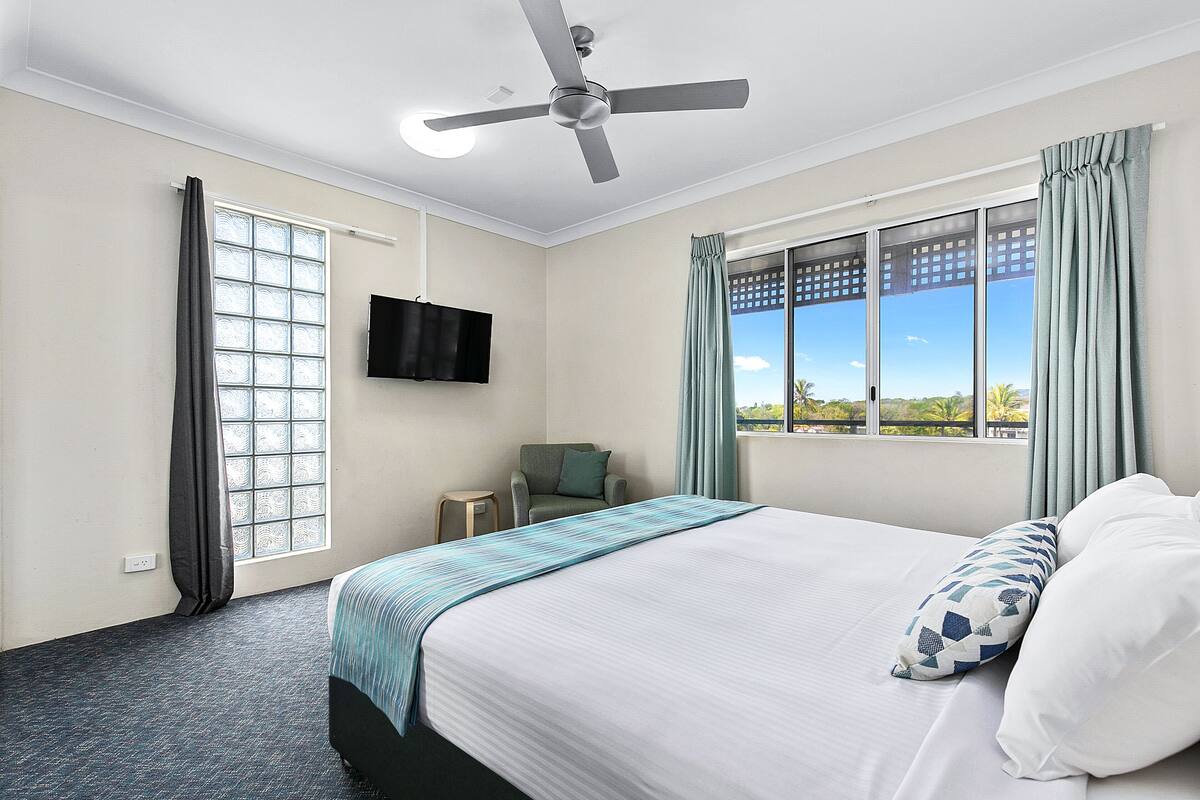 A hotel room with a bed, a ceiling fan, a TV, a chair, and a window with curtains.