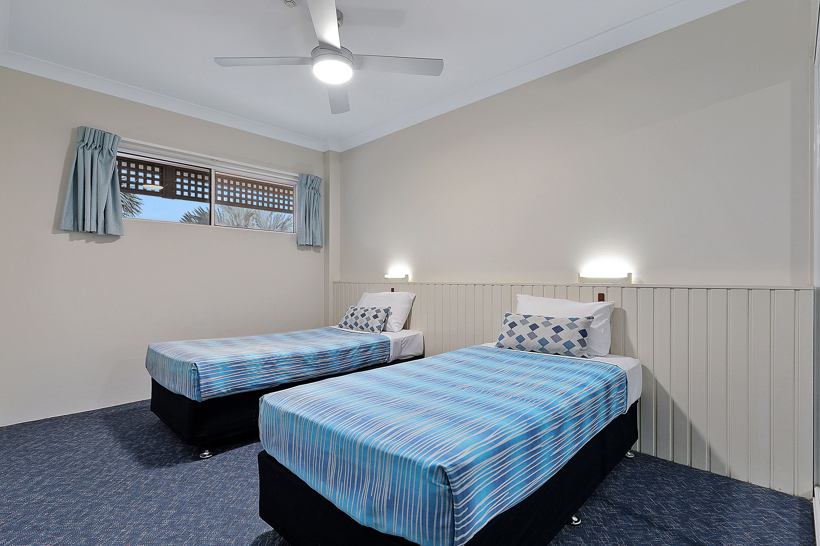 Una habitación con dos camas individuales, un ventilador de techo y una ventana con cortinas.