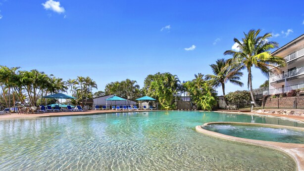 A clear pool with lounge chairs and umbrellas, surrounded by palm trees and a building with balconies. - K'gari Beach Resort (Eurong)