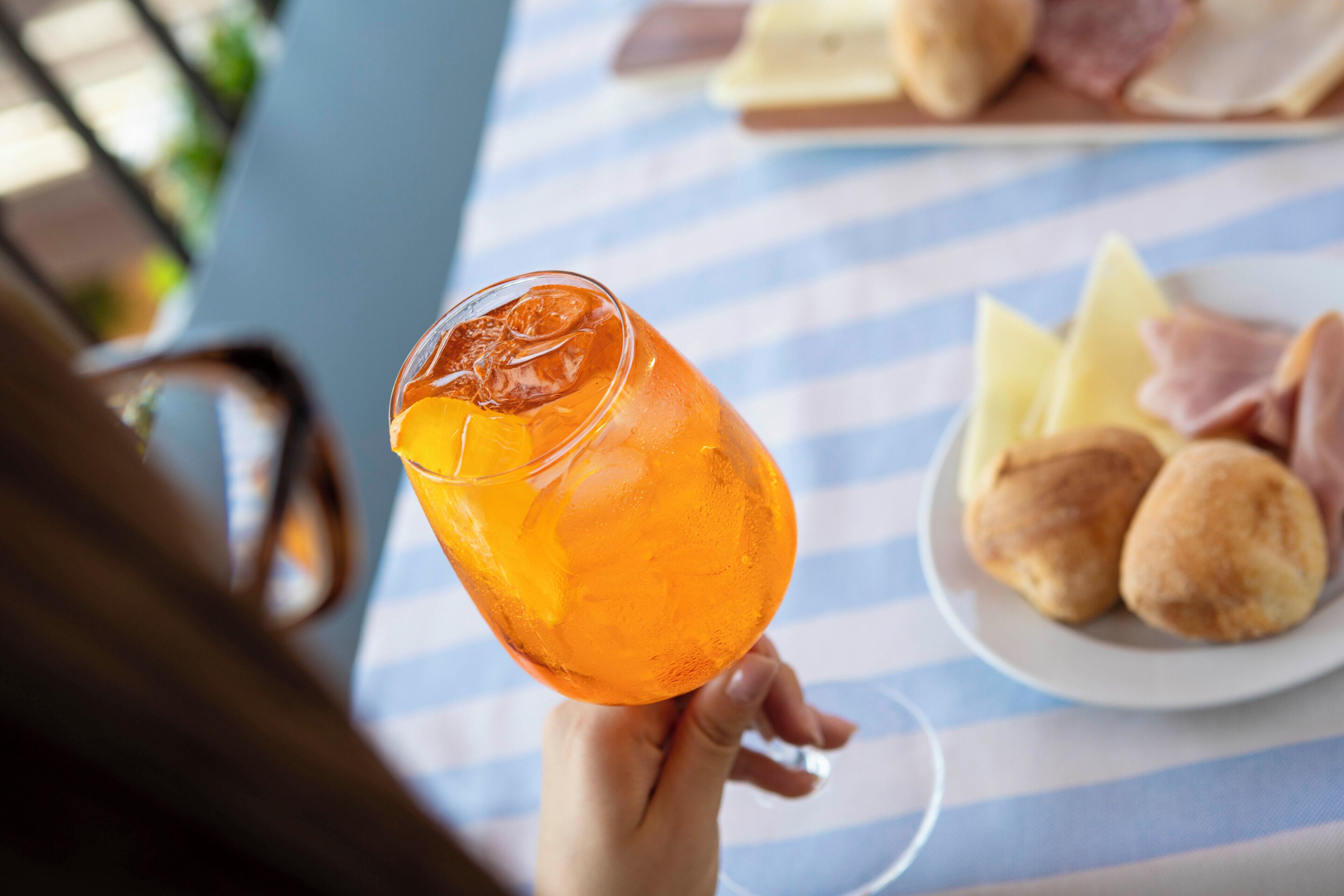 Una persona sosteniendo un vaso de bebida de naranja con cubitos de hielo, un plato de queso y panecillos, y un mantel a rayas.