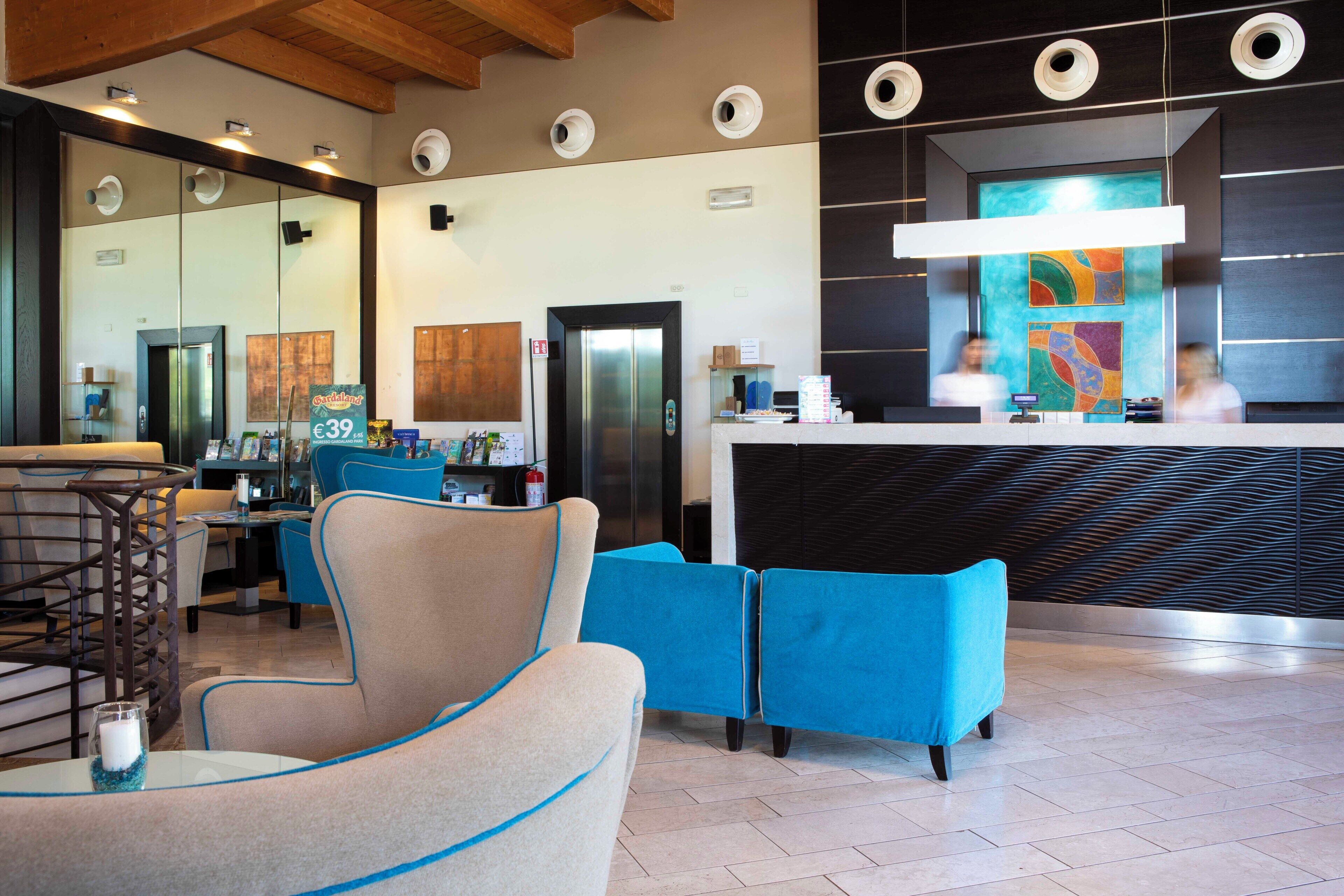A modern hotel lobby with a reception desk, seating area, and a large mirror reflecting the space.