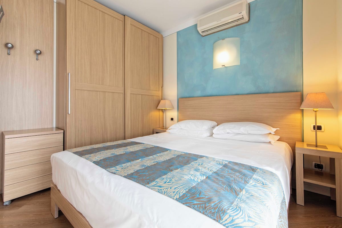 Habitación de hotel con una cama grande, armario de madera, mesitas de noche con lámparas y un equipo de aire acondicionado en la pared.