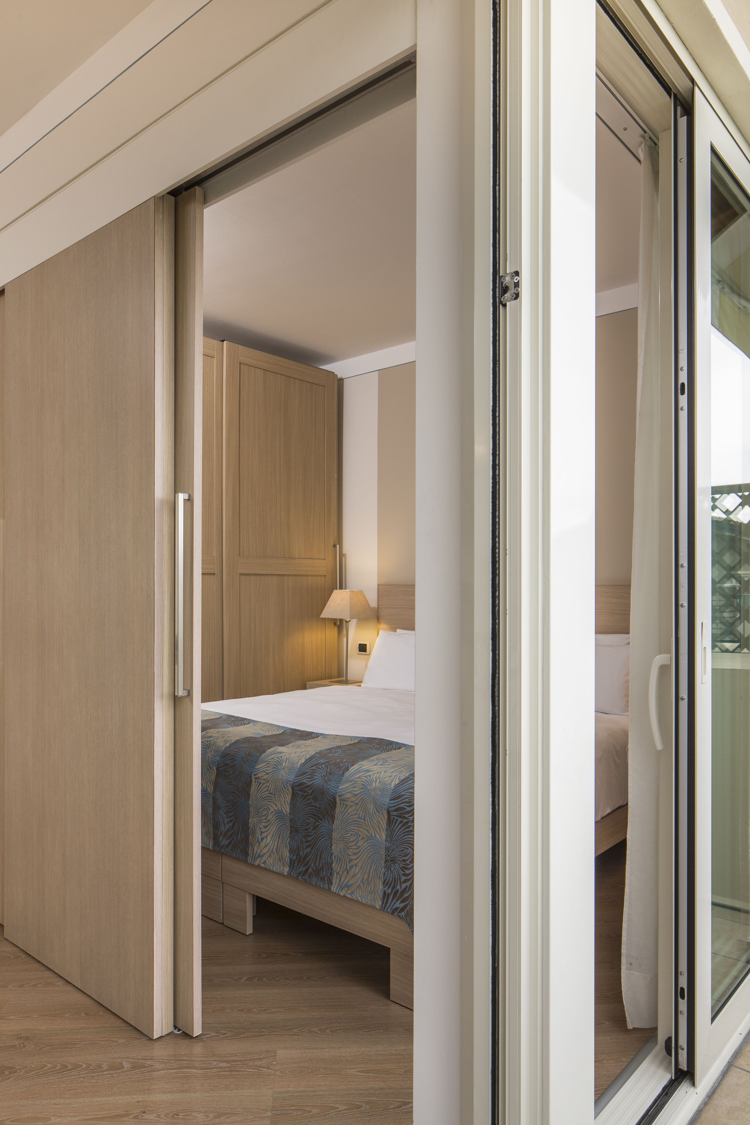 Habitación de hotel con cama, mesita de noche, lámpara y puerta corrediza que da a un balcón.