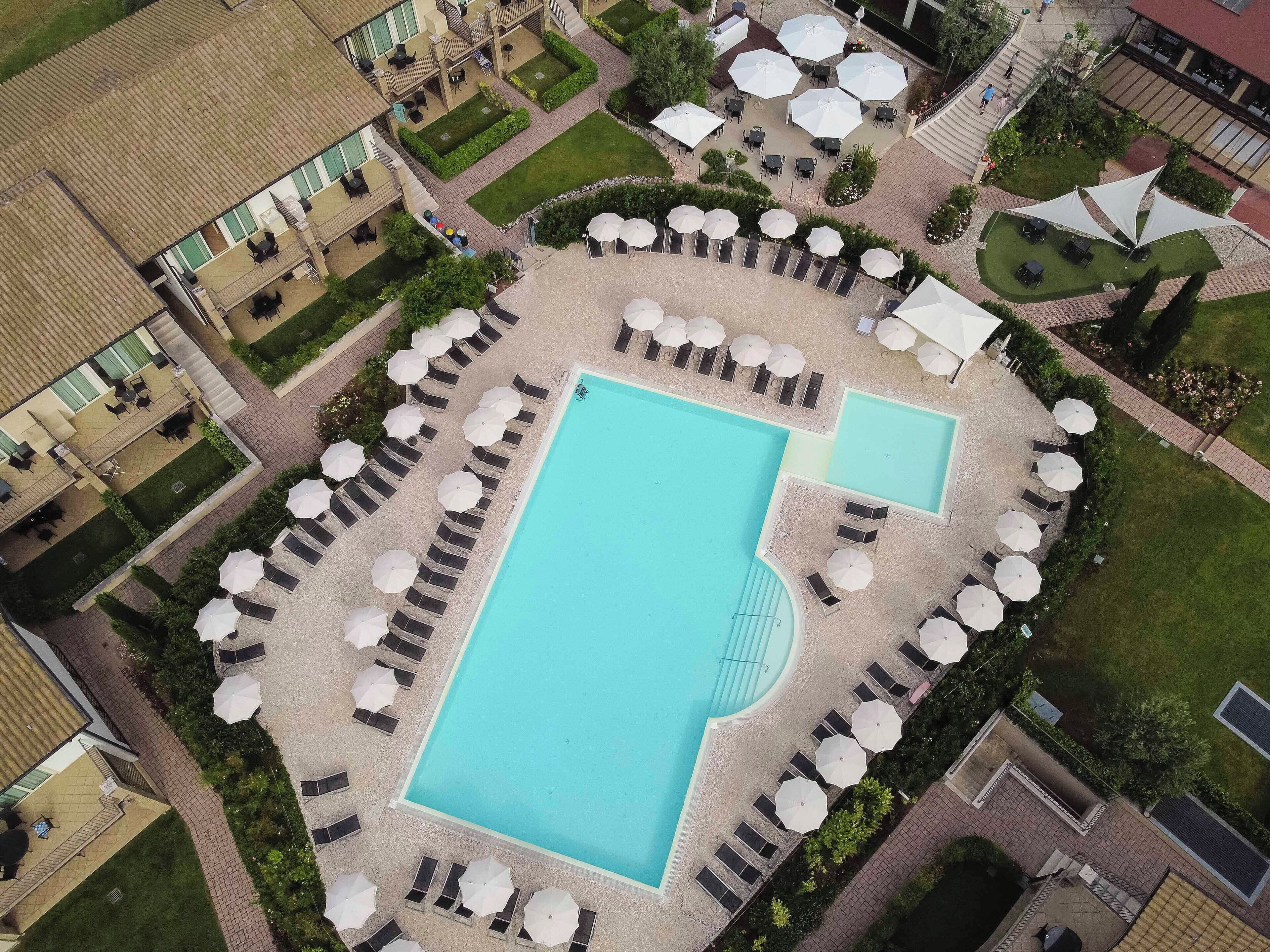 Vue aérienne d’un hôtel doté d’une grande piscine centrale entourée de transats et de parasols, ainsi que d’une plus petite piscine située à droite.