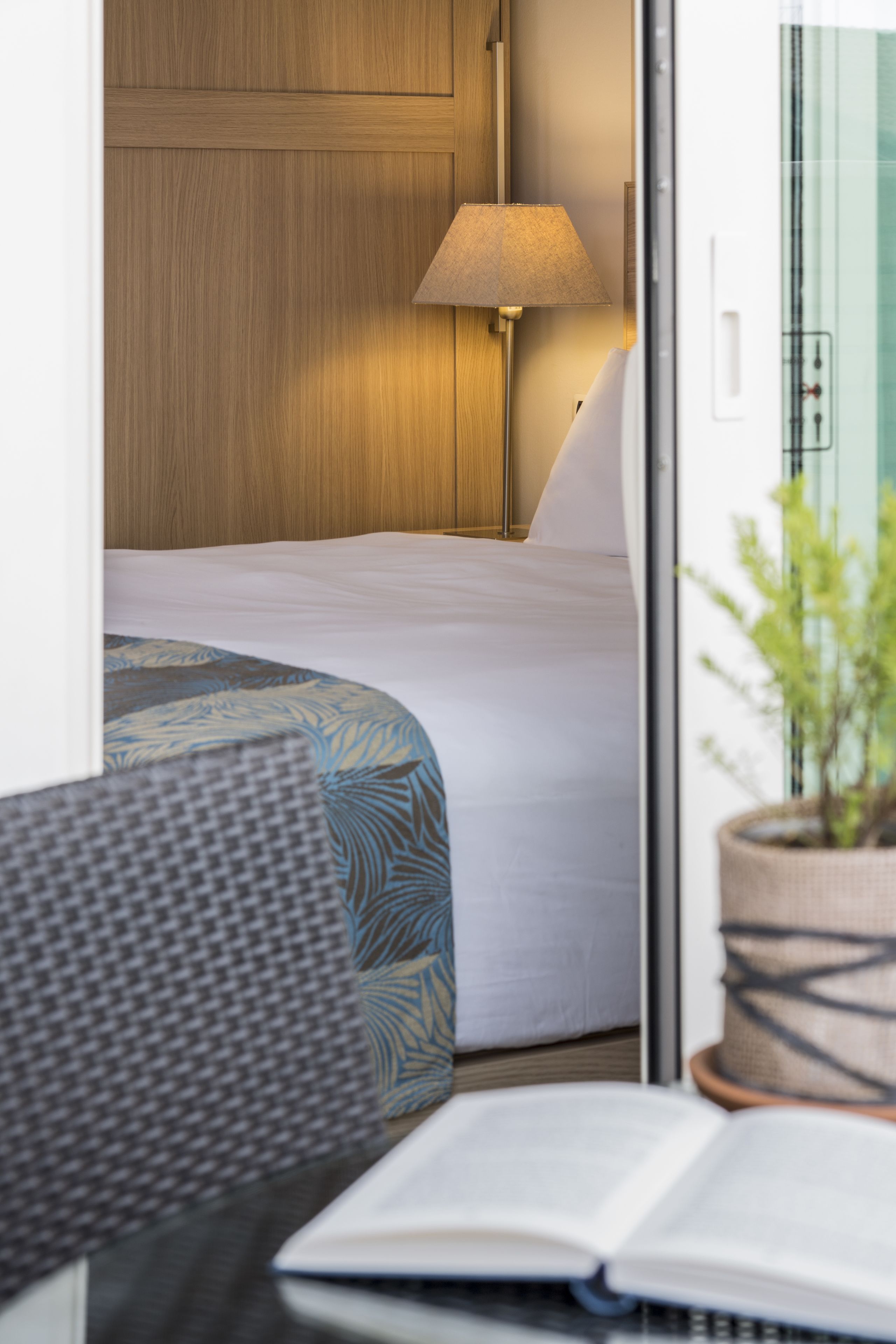 Una habitación de hotel con cama, lámpara, planta en maceta y un libro sobre una mesa.