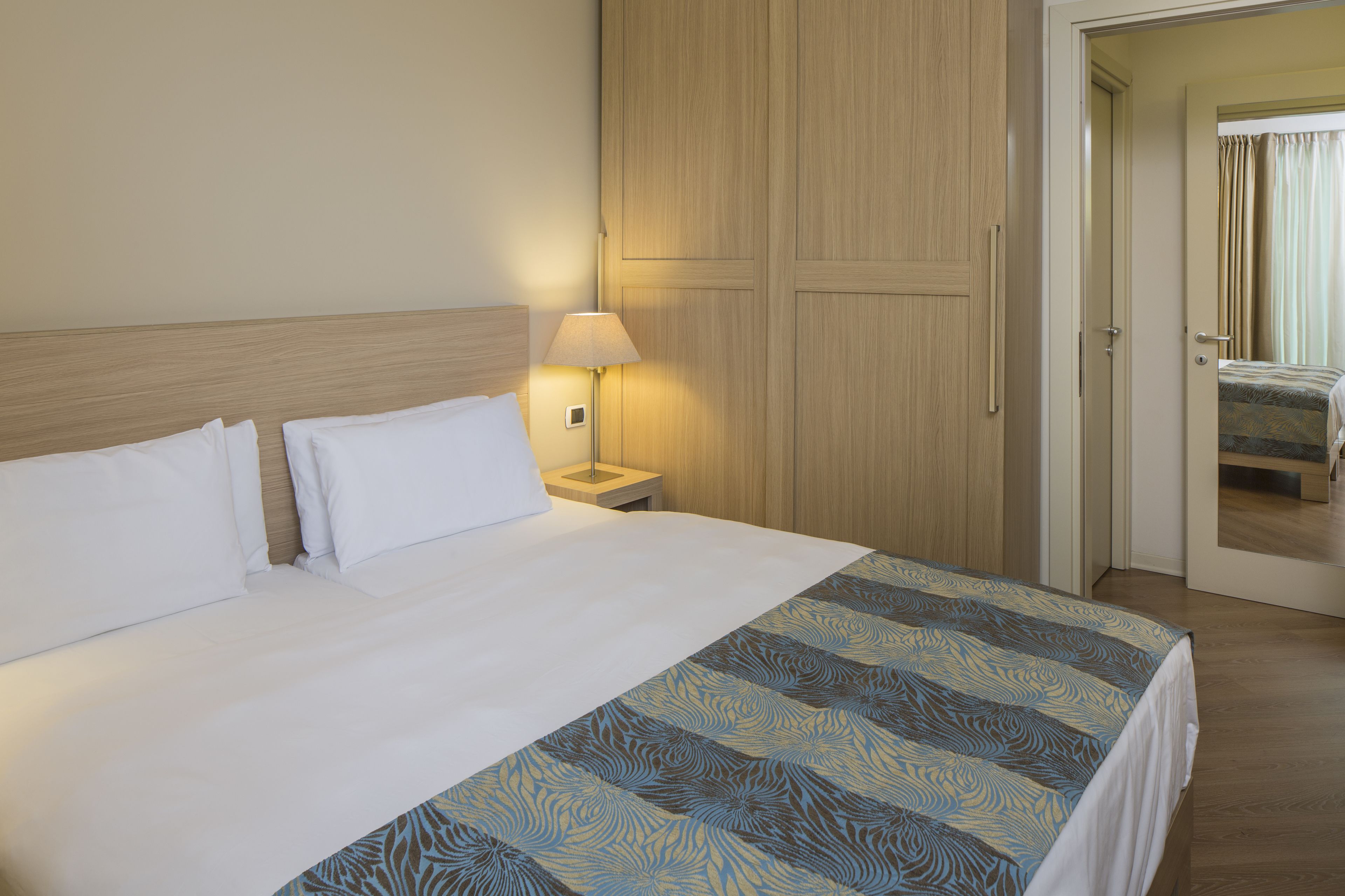 A hotel room with a large bed, a nightstand with a lamp, wooden wardrobe, and a door leading to another room.