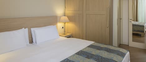 A hotel room with a large bed, a nightstand with a lamp, wooden wardrobe, and a door leading to another room.