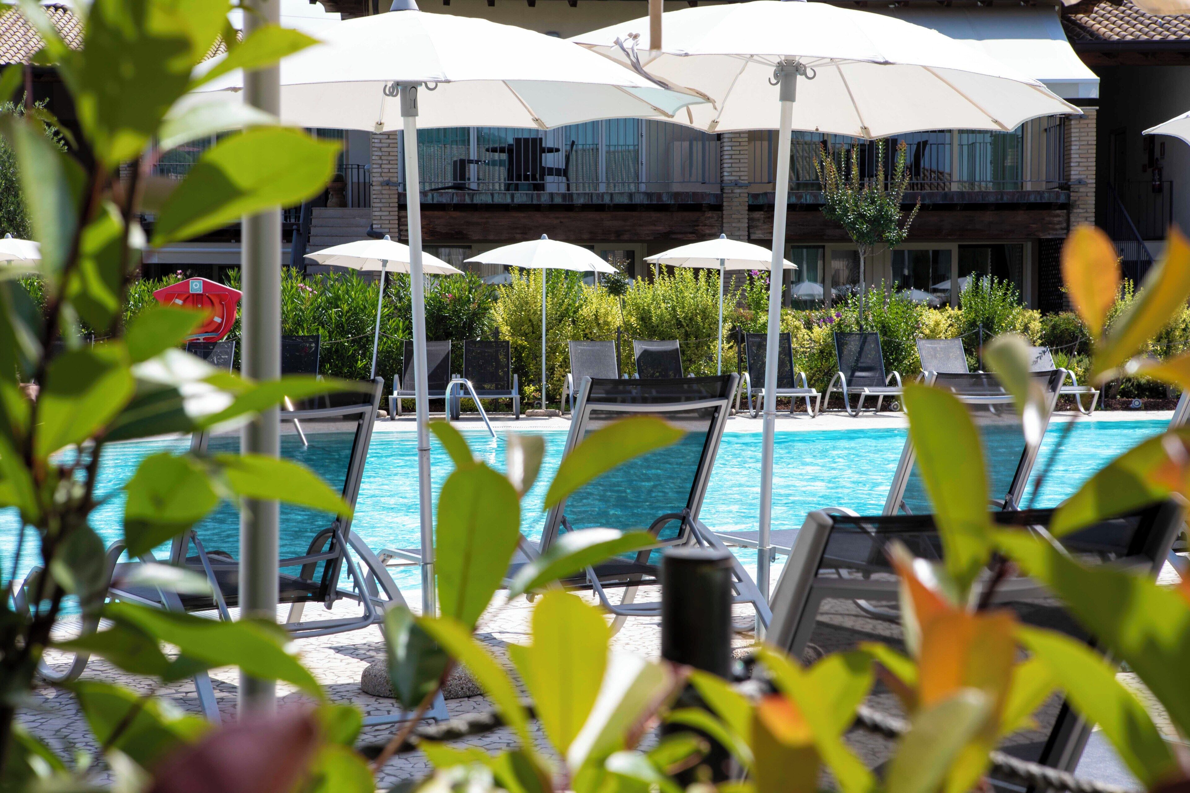 Un área junto a la piscina con sillones de descanso y sombrillas, una piscina cristalina y un edificio con balcones al fondo.
