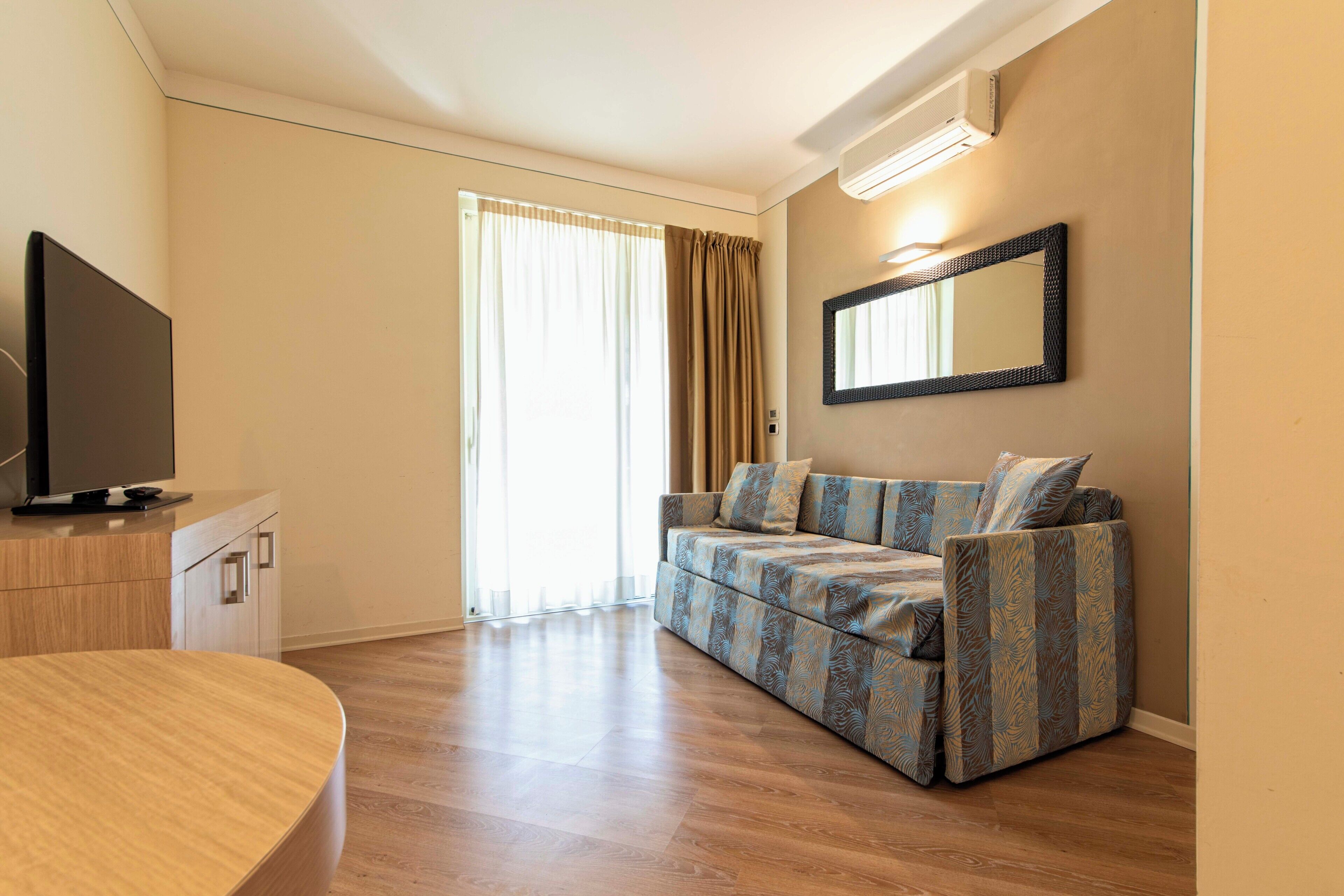 A modern living room with a flat-screen TV, a sofa, a dining table, and a large window with curtains.