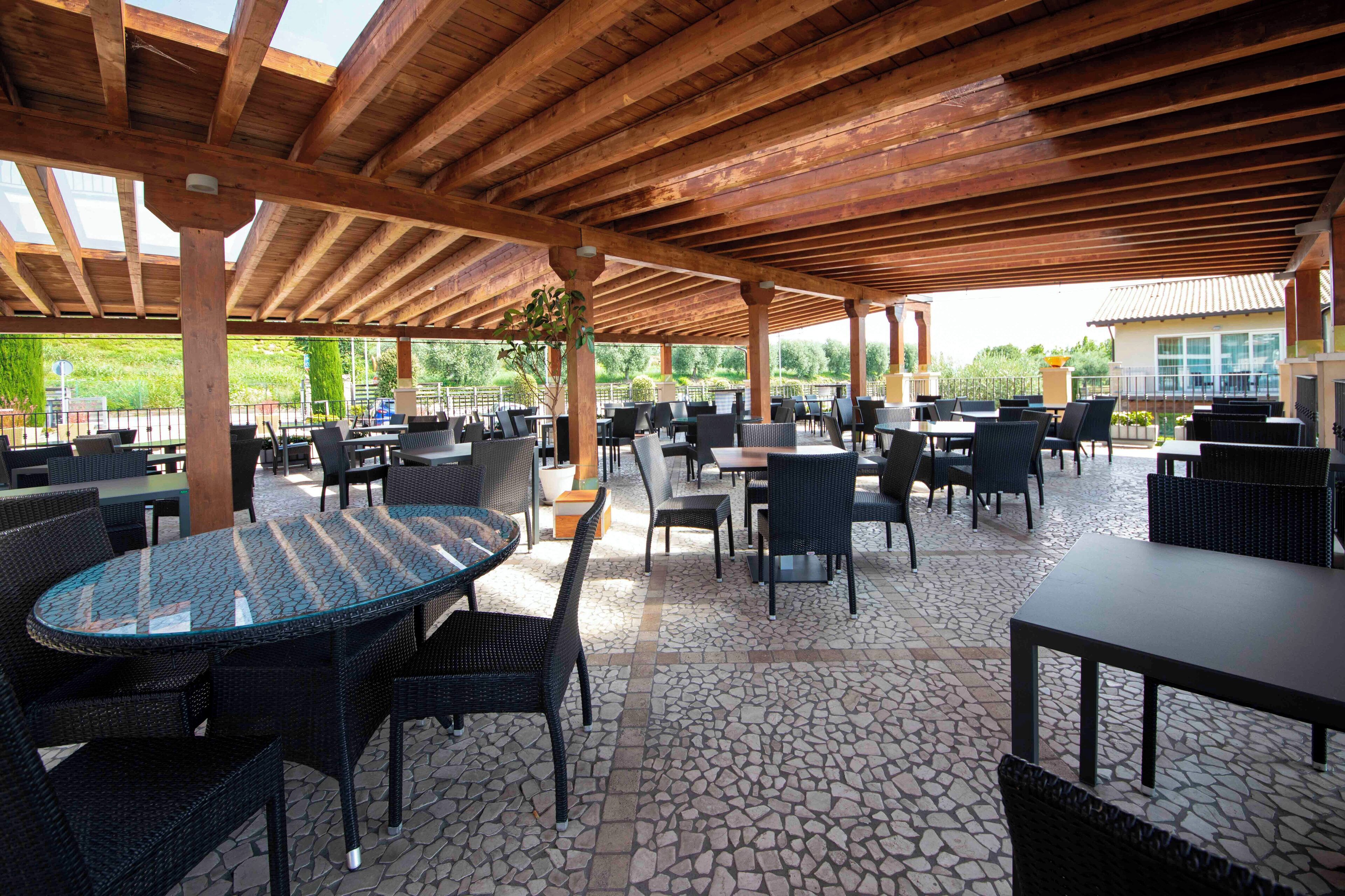 Uma área de pátio ao ar livre com várias mesas e cadeiras, vigas de madeira no teto e piso de ladrilho de pedra.