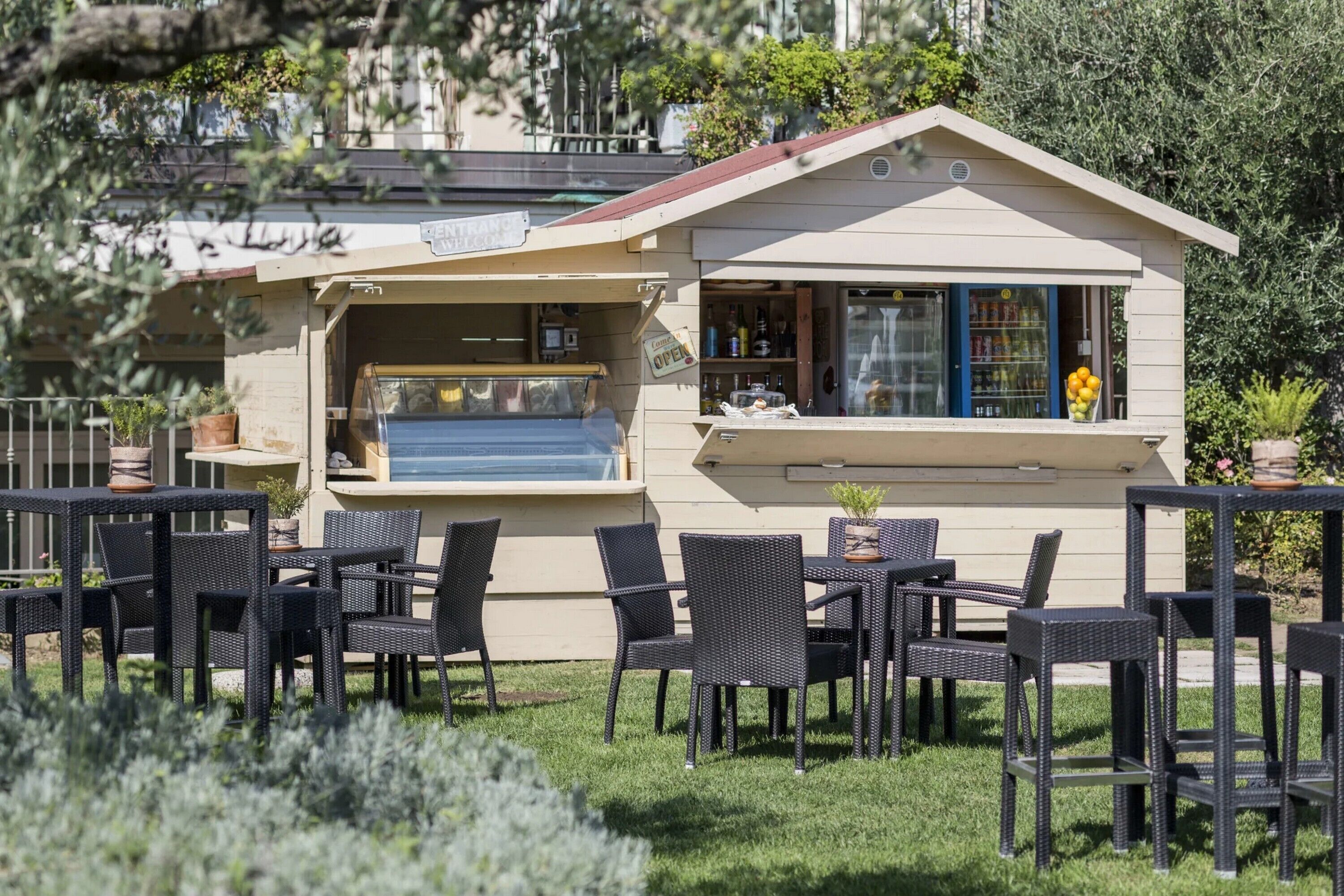 Un área de estar al aire libre con sillas y mesas de mimbre, una zona de bar con botellas y un letrero, y un edificio con fachada color beige y techo rojo.