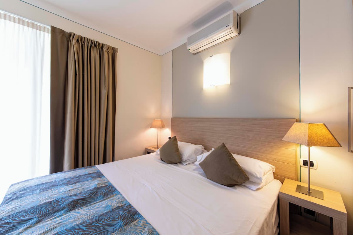 Habitación de hotel con cama, cortinas, aire acondicionado, mesita de noche con lámpara y luz empotrada en la pared.