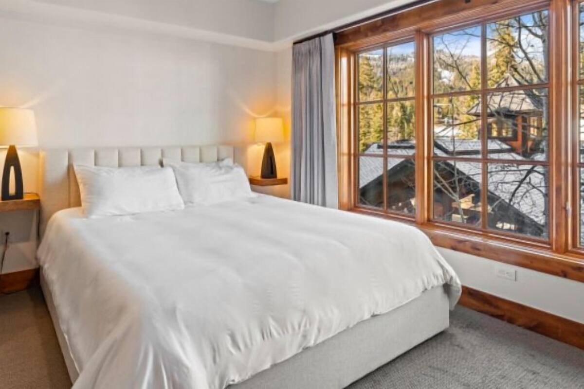 A hotel room with a bed, two bedside lamps, a chair, and a large window with a view of trees and a building.