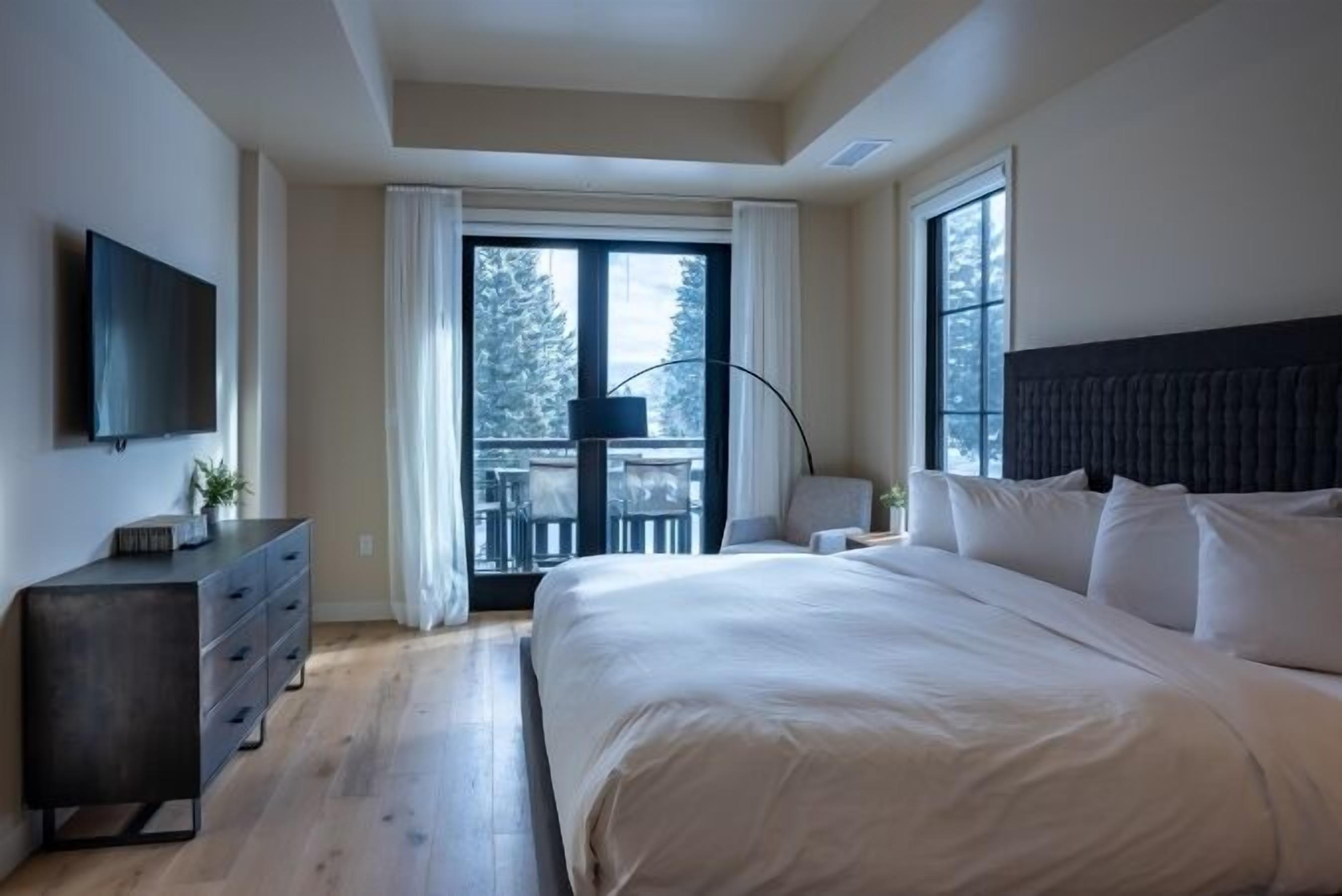 Un dormitorio con una cama grande, un televisor en la pared, un aparador y un balcón con vistas a árboles.
