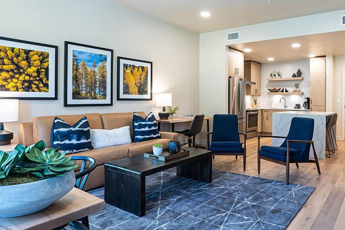 A modern living room with a sofa, coffee table, and chairs. There is a kitchen area with a refrigerator, stove, and sink.