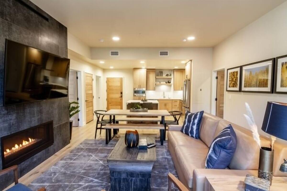 A modern living room with a fireplace, a sofa, a dining table, and a kitchen in the background.