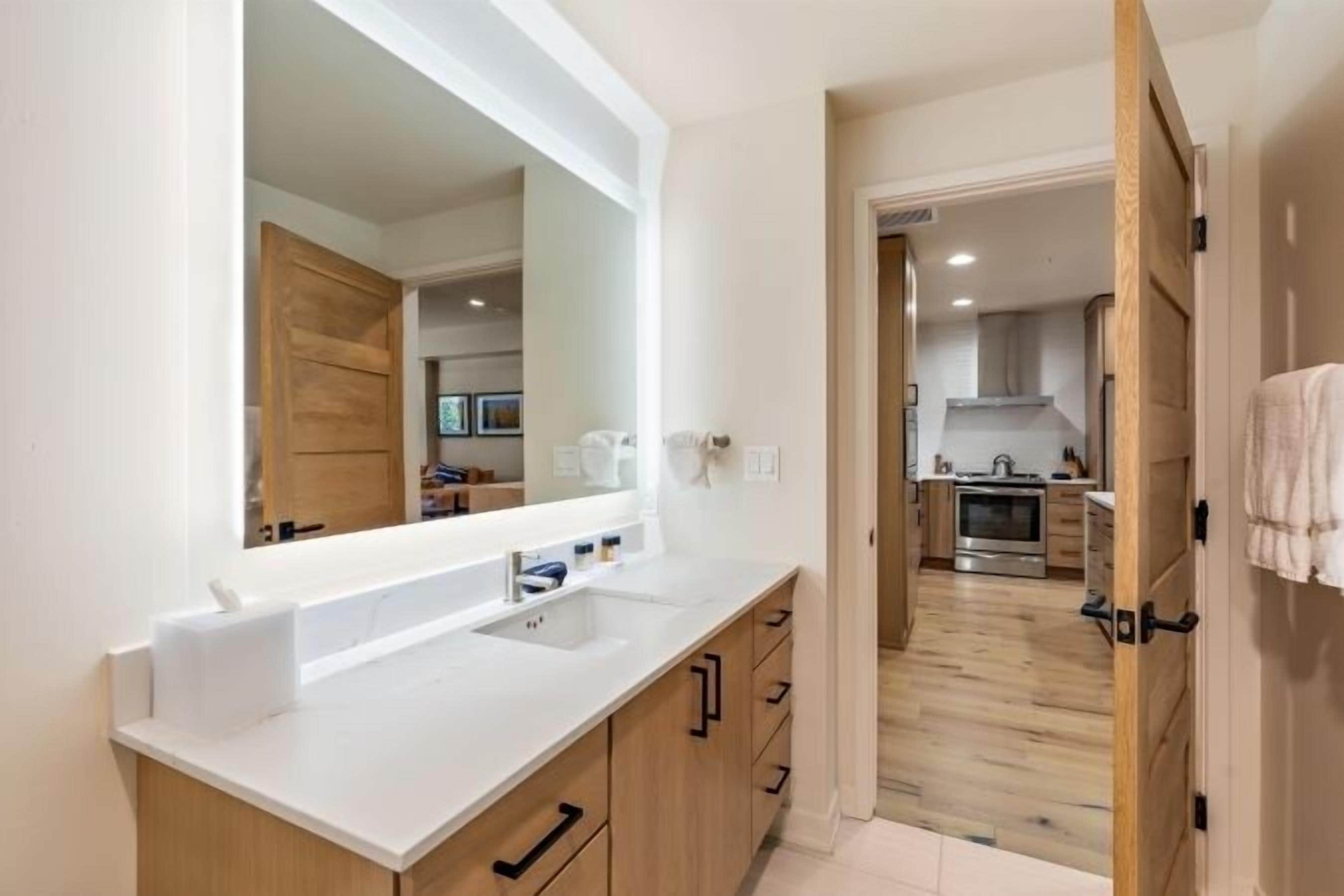Une salle de bain moderne avec un grand miroir, un plan de travail blanc, des meubles en bois et une vue sur la cuisine.