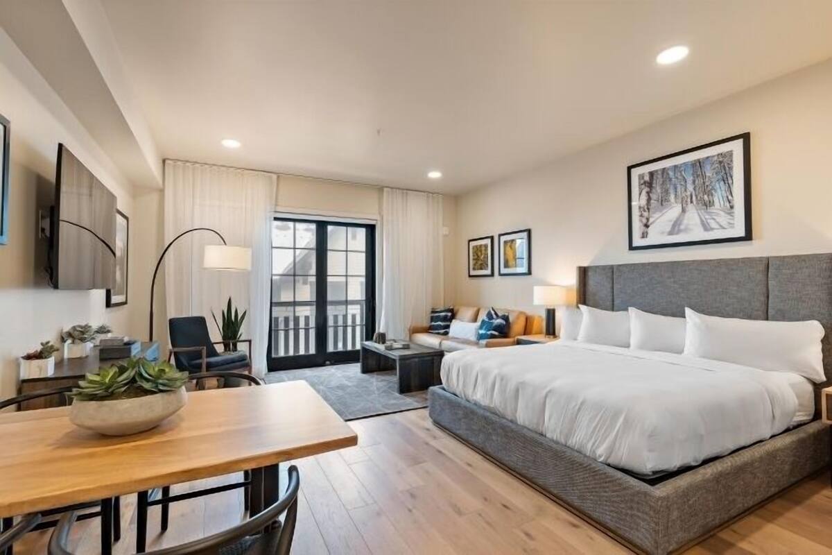 A hotel room with a large bed, a desk, a chair, a sofa, and a balcony with curtains.