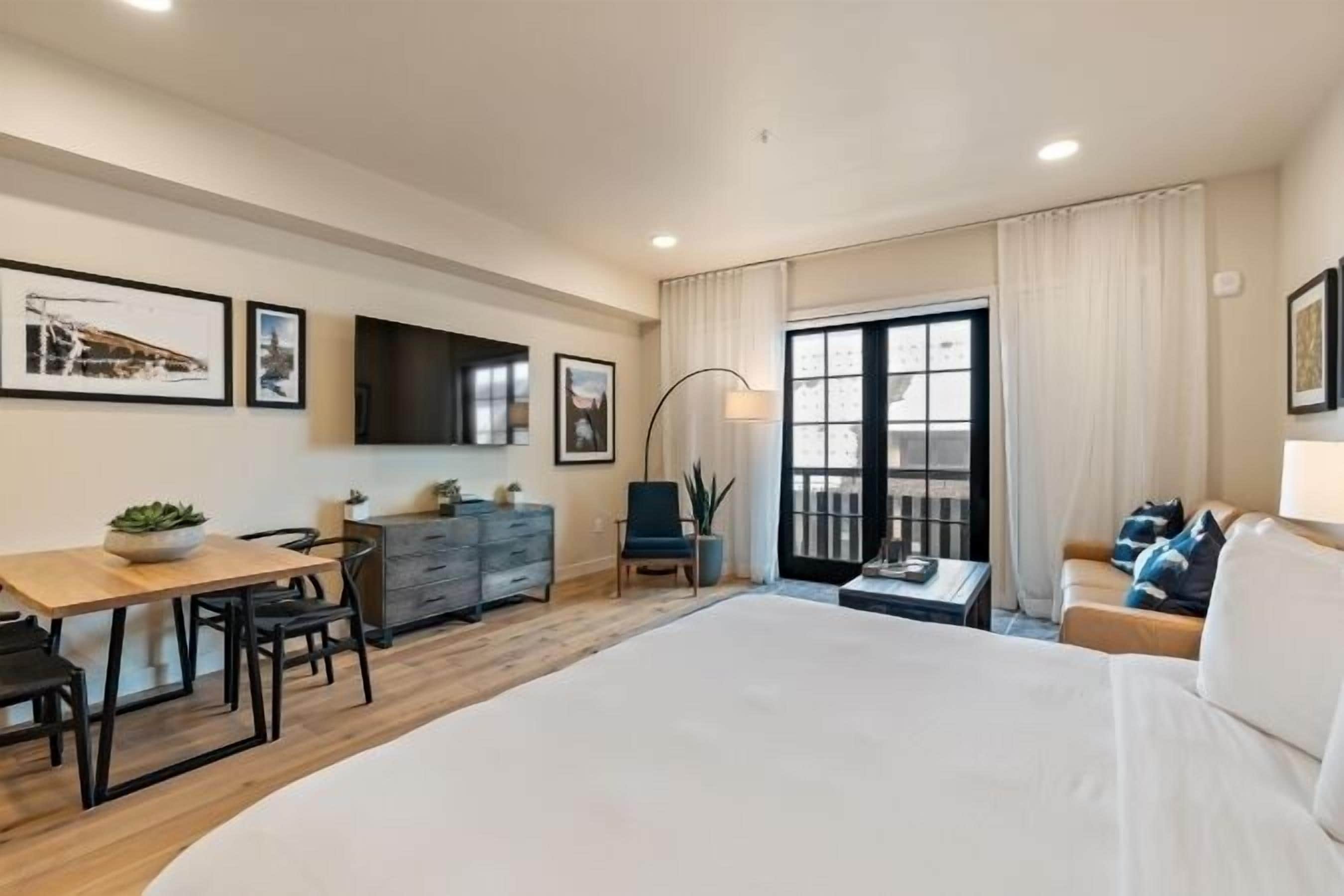 A hotel room with a large bed, a desk with chairs, a TV mounted on the wall, and a balcony with curtains.