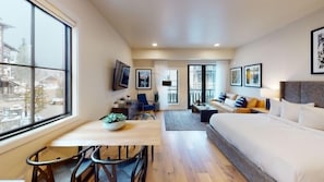 A compact hotel room with a bed, a desk, and a chair. There are framed pictures on the walls and a window with a view of buildings.