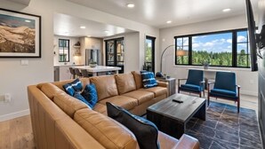 A modern living room with a leather sofa, a coffee table, and blue chairs. There is a dining area with a table and chairs in the background.