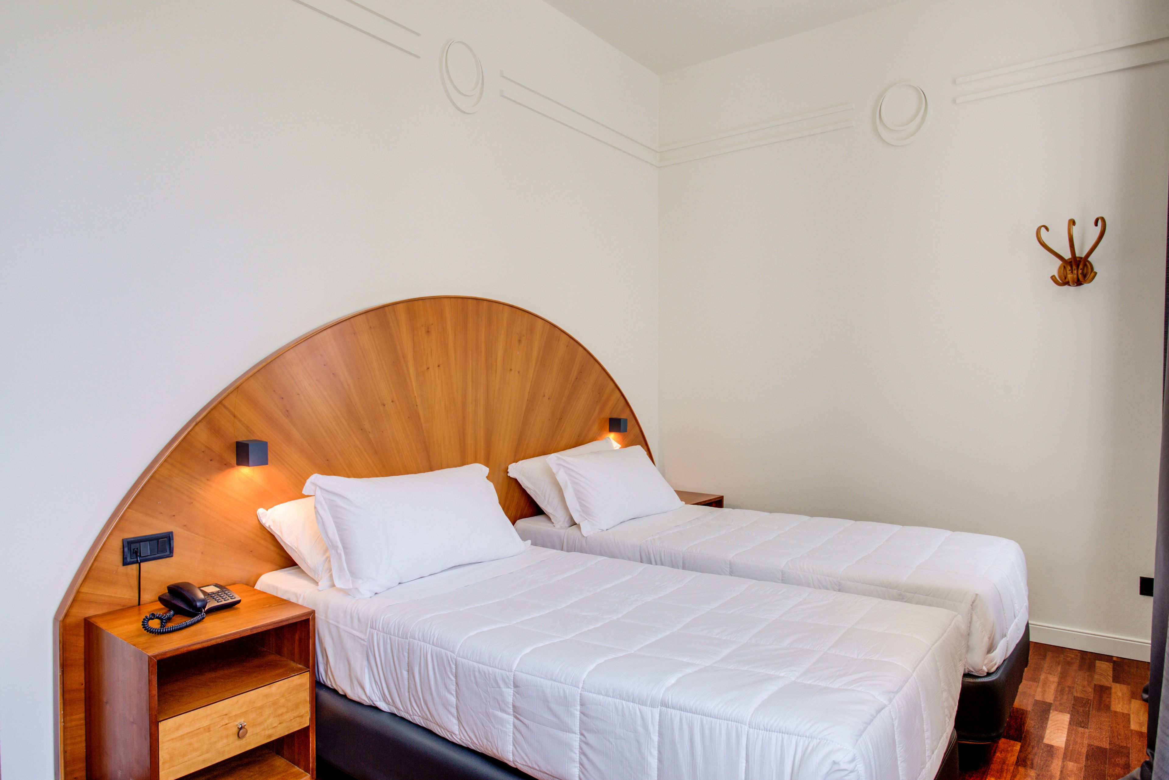A room with two beds, a wooden headboard, a bedside table with a phone, and a wall-mounted hook.