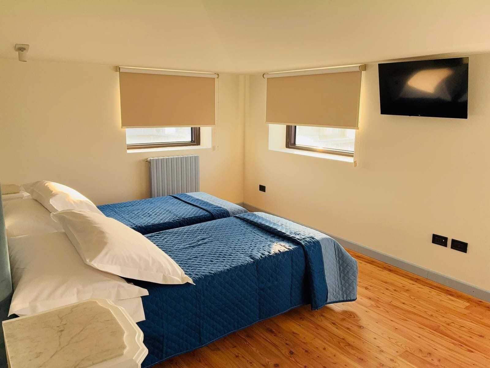 A bedroom with a bed, two windows with blinds, a radiator, and a TV on the wall.