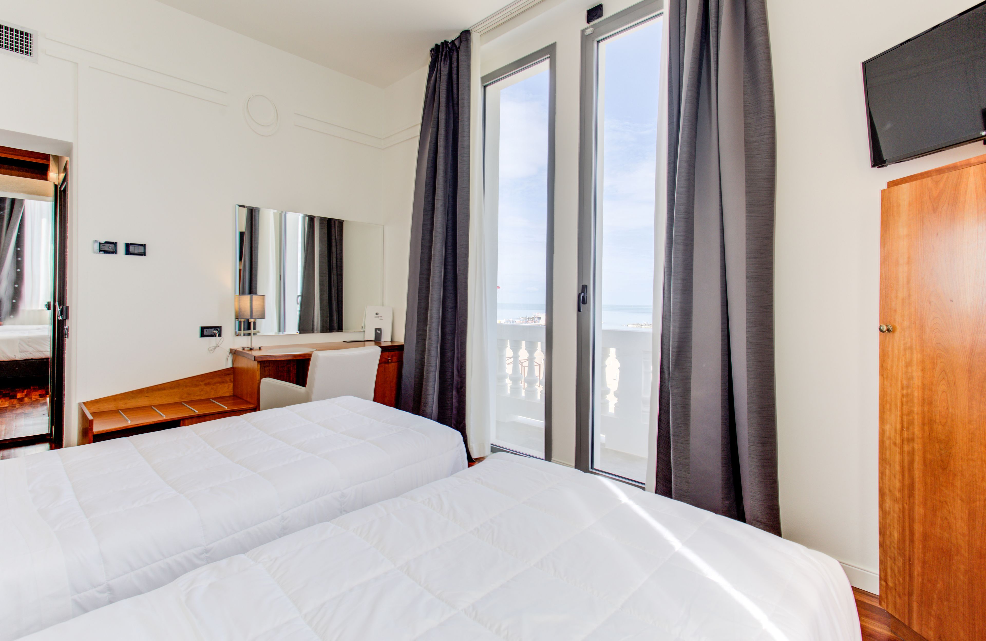 A hotel room with two beds, a desk, a chair, a TV, and a balcony with curtains.