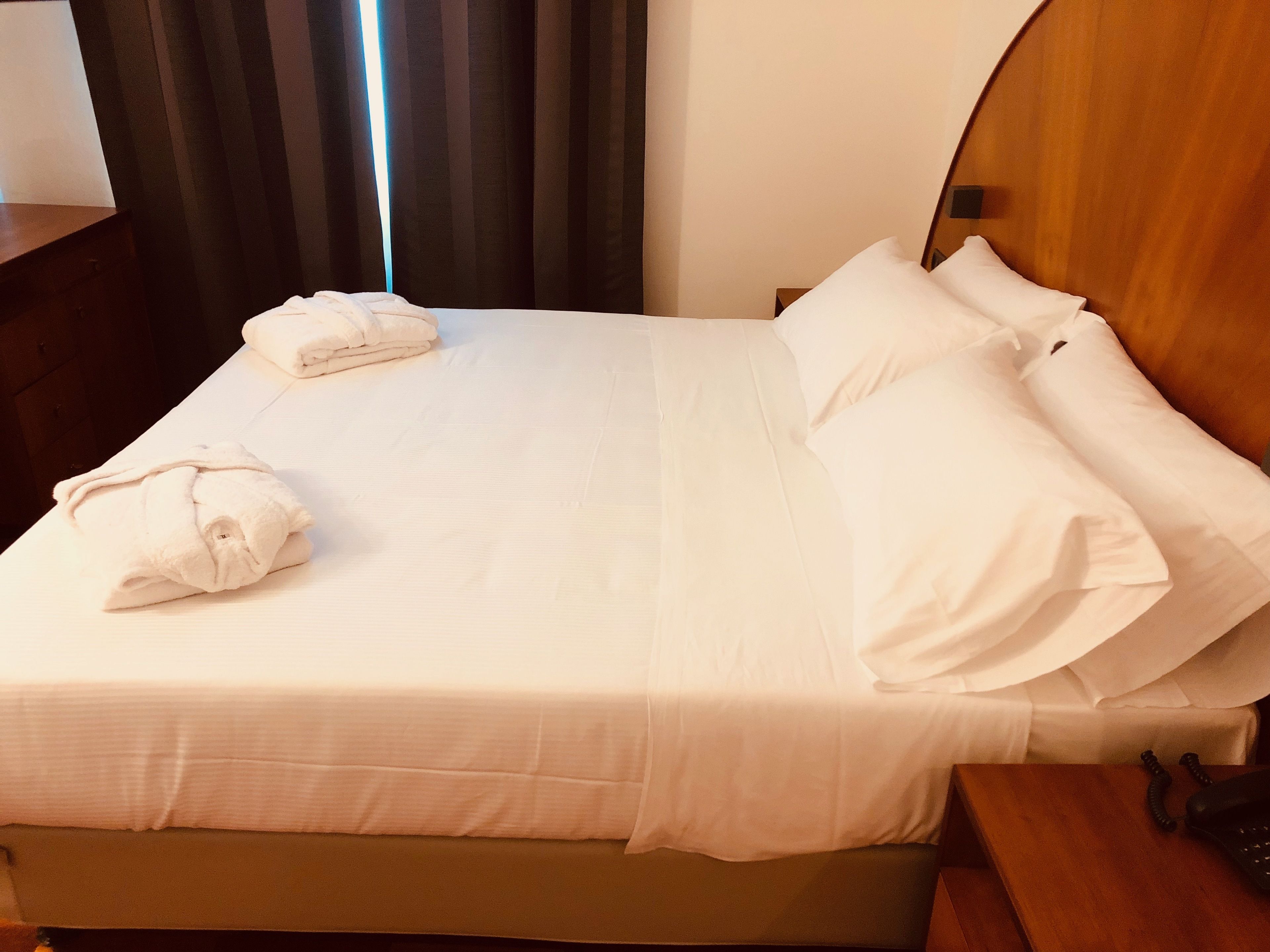 A neatly made bed with white linens and pillows, a folded towel, and a wooden headboard.