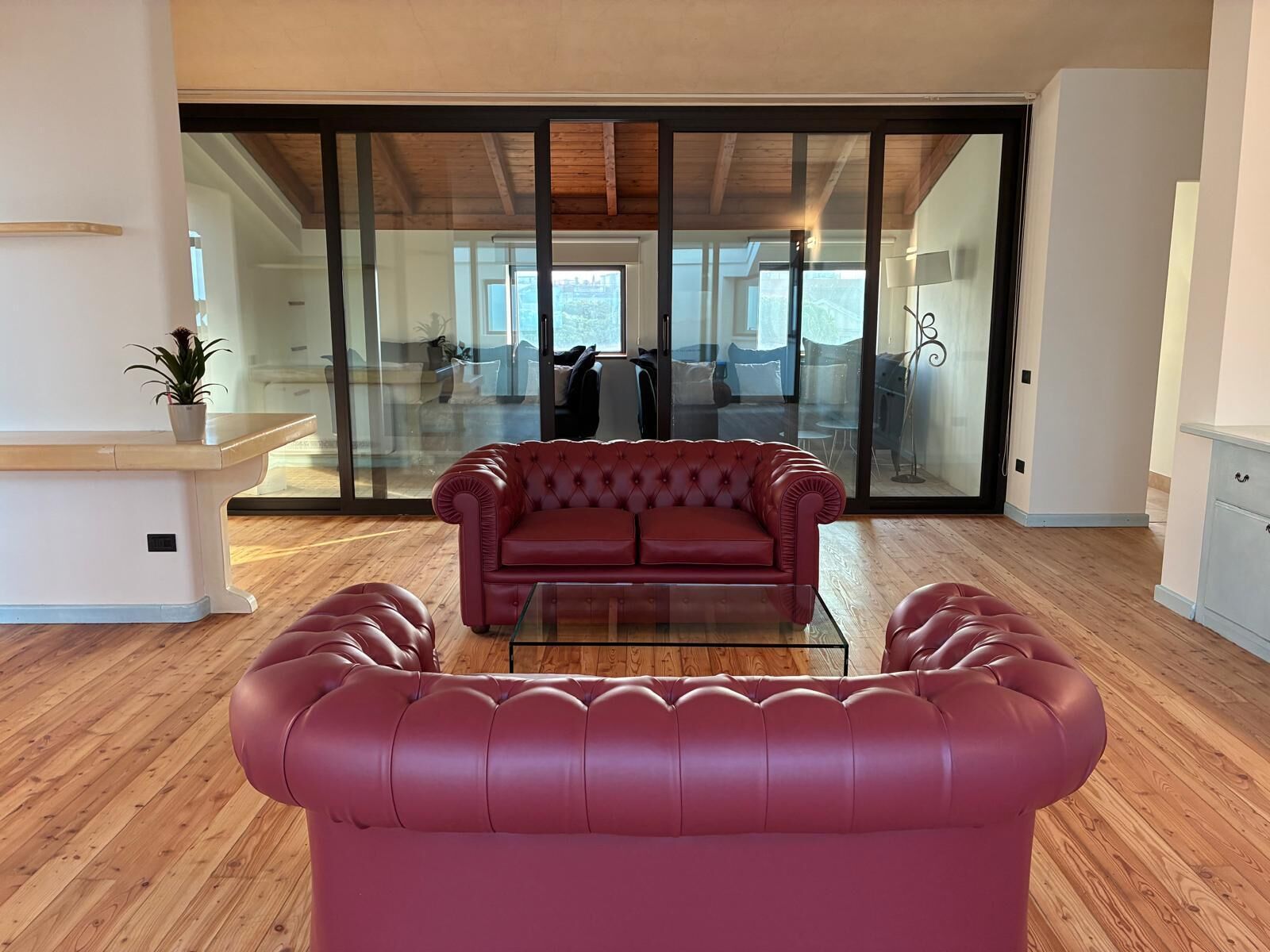 Uma sala de estar moderna com um sofá de couro vermelho, uma mesa de centro de vidro, piso de madeira e uma porta de vidro deslizante que dá para uma varanda.
