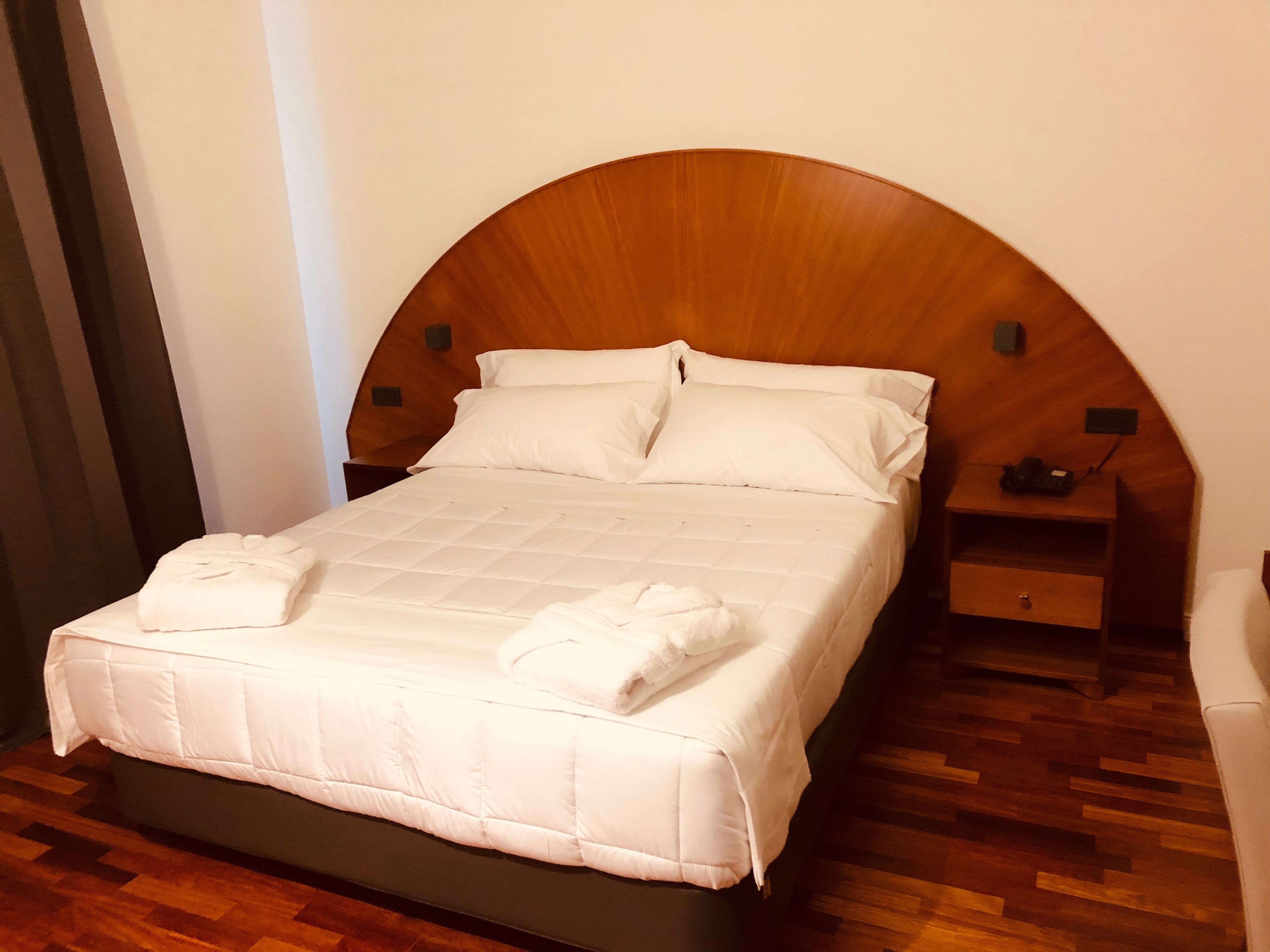 Uma cama bem arrumada com lençóis brancos e duas toalhas, uma cabeceira de madeira, uma mesa de cabeceira com um telefone e piso de madeira.