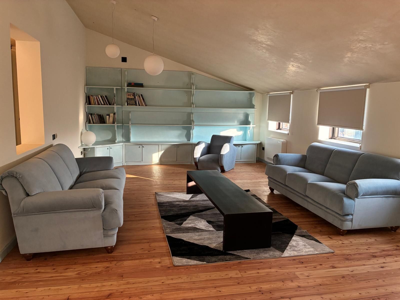 A spacious living room with a high ceiling, a built-in bookshelf, two grey sofas, a black coffee table, and a patterned rug.