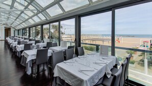 A restaurant with multiple tables set for dining, large windows offering a view of the beach and ocean, and a modern architectural design with a glass ceiling. - Kursaal Hotel (Cattolica)
