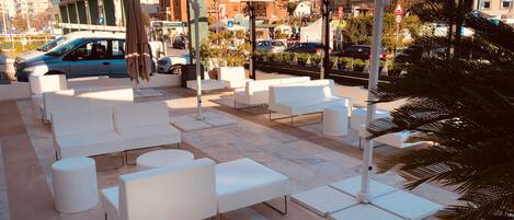 Outdoor seating with white sofas and round tables, umbrellas, and a cityscape background.