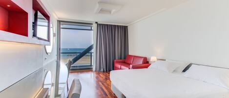 Habitación de hotel moderna con una cama grande, un sofá de cuero rojo y vista al océano a través de un ventanal amplio.